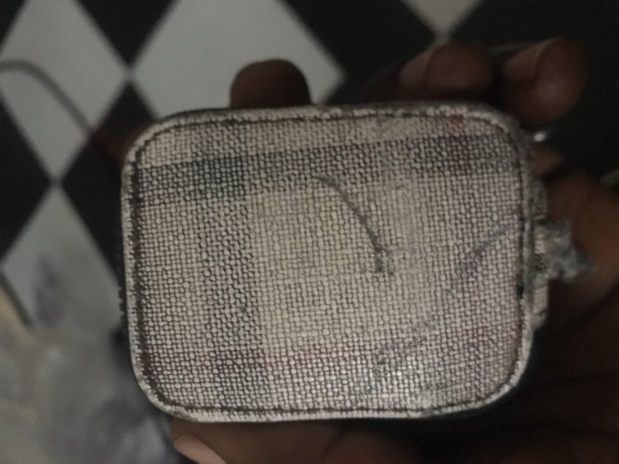A close-up, top-down shot shows a hand holding a small, rectangular object. The object is covered in a coarse, white fabric with a grid-like pattern of tiny dots. The object has rounded corners and appears to be worn, with some dark marks and a slight fraying of the fabric along the edges. A faint curved line is visible on the surface of the object. The background is blurred, showing a black and white checkered pattern, suggesting an indoor environment.
