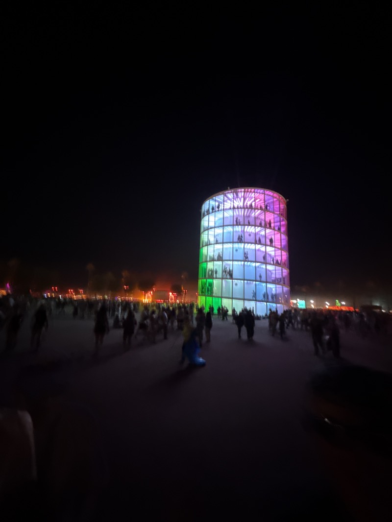 The image shows a large, cylindrical structure illuminated with colored lights, possibly a building or art installation. The structure is multicolored, with green at the bottom, transitioning to blue, then pink at the top. People can be seen on different levels of the structure.
The image appears to be taken at night, in an outdoor area with many people. The crowd is a mix of people standing, walking, and interacting. There are other light sources and structures in the background, but they are less distinct.
The location is in the city of Indio, United States, likely at the Coachella Music Festival.
