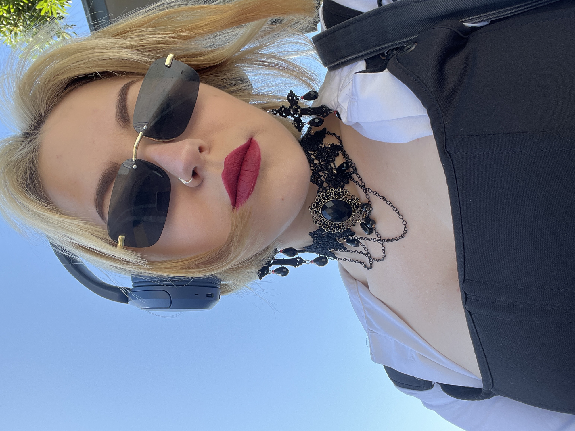 A low-angle shot depicts a woman with blonde hair and red lipstick, wearing dark sunglasses and headphones. She has a nose ring and a black necklace with an ornate pendant. Her clothing appears to consist of a white shirt with a black vest or overall-type garment. The background is a bright blue sky, with green foliage visible in the top left corner. The location cannot be confirmed as Bečići, Montenegro, from the available visual information.