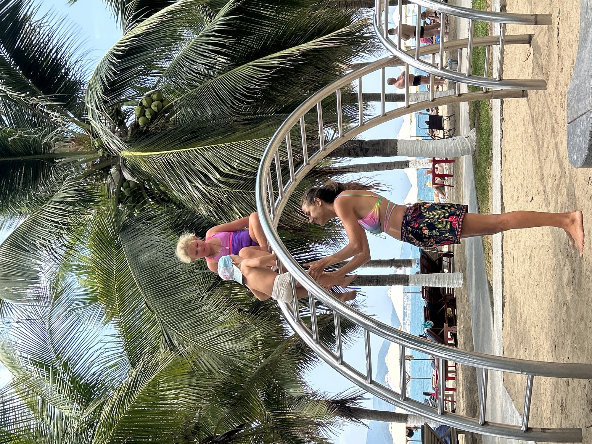 The image shows a family interacting with a metal climbing structure on a beach. A woman is standing on the ground, her hands extended toward a child who is climbing the structure. An older woman is already on the structure, seemingly supervising the child. The location appears to be a beach in Nha Trang Ward, Vietnam, as suggested by the coconut trees, sandy beach, and the presence of other people on the beach in the background. Beach chairs and umbrellas are visible along the shore. The sky is clear and blue, suggesting a sunny day.