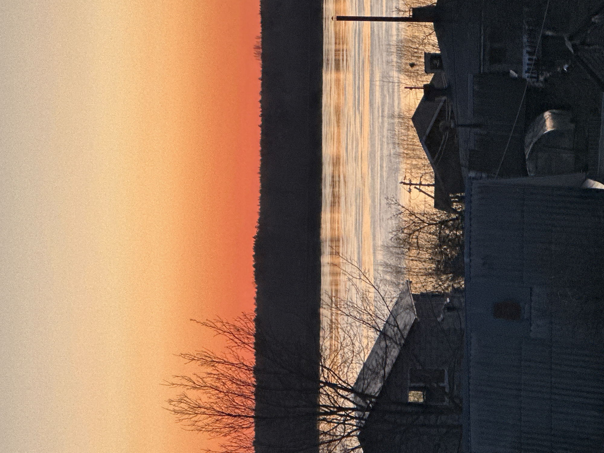 The image shows a view of a river at sunset in the city of Chaikovsky, Russia. Silhouetted trees and buildings are visible on the right side of the frame, with their outlines against the background of the river and the orange and yellow sky. The river reflects the light of the setting sun. The landscape consists of a forest, body of water, and structures on the horizon. The scene evokes a quiet evening or early morning.