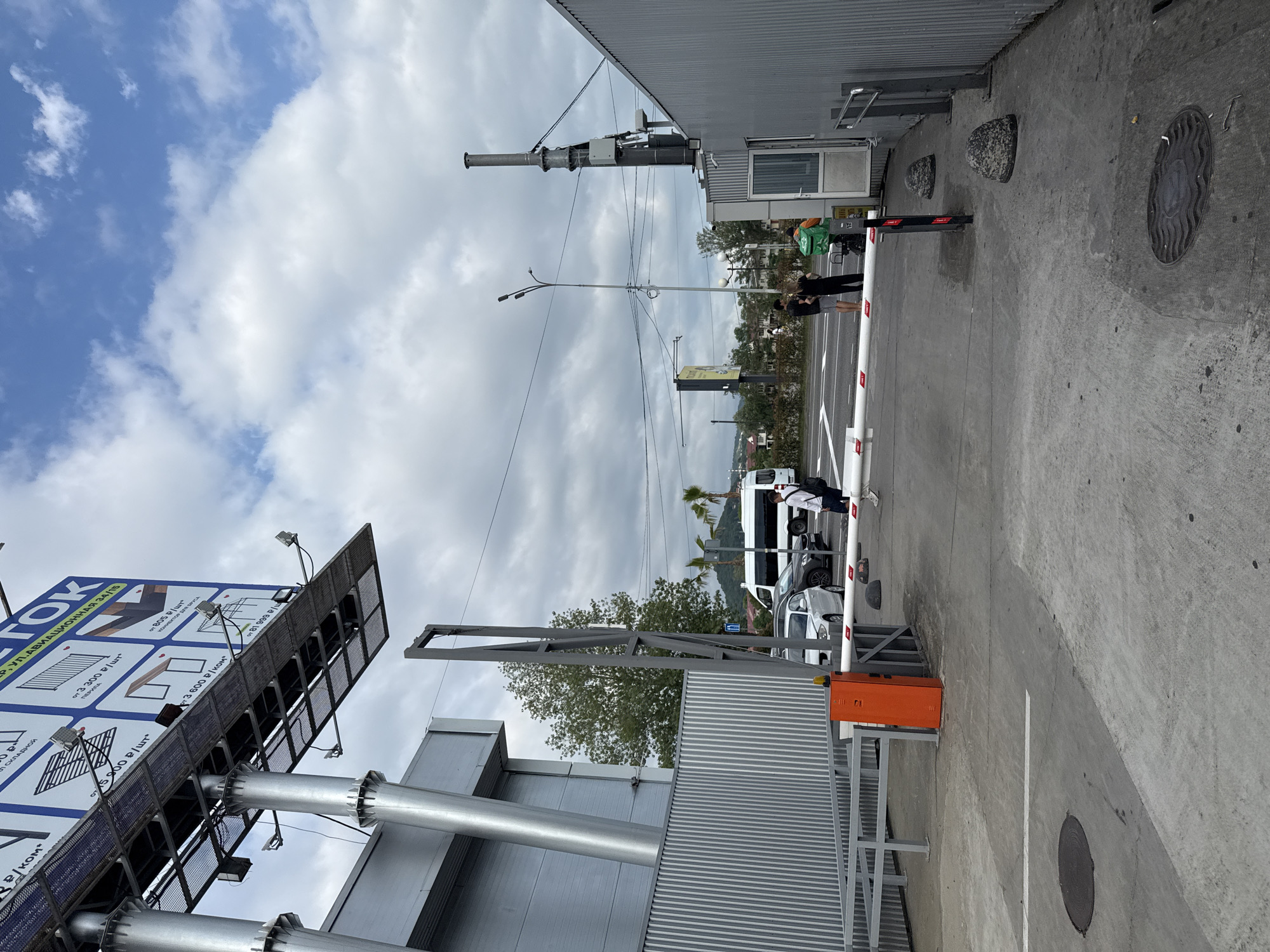 This media file captures a scene at what appears to be an entrance or checkpoint, possibly for a business or parking area. The perspective is from ground level, looking towards the sky and across a paved area.
In the foreground, a metal barrier arm is raised, indicating passage is allowed. An orange control box is visible at the base of the barrier. The ground is concrete, with painted white lines marking a roadway. Several cars are visible beyond the barrier, including a white van and a dark sedan.
To the left of the scene, a large billboard is prominently featured, displaying advertisements. The text on the billboard, partially visible, includes Cyrillic script and mentions prices in rubles, with phrases like "складной" (folding) and "перила" (railings). This suggests the billboard is advertising construction materials or home improvement products.
Further into the background, there are structures that look like buildings, possibly commercial or industrial, with corrugated metal siding. A security booth or small building is also visible.
Two people are standing near the security booth, one appearing to wear an orange vest, possibly a security guard or attendant. Another person is walking away from the barrier. The sky is partly cloudy, with a mix of blue sky and white clouds, suggesting it is daytime, likely late morning or afternoon. The weather appears overcast or intermittently sunny. In the distance, trees and some hilly terrain are visible, hinting at a location with some natural landscape features. The presence of palm trees in the background could suggest a warmer climate. The overall atmosphere seems functional and urban, with elements of outdoor advertising and access control.