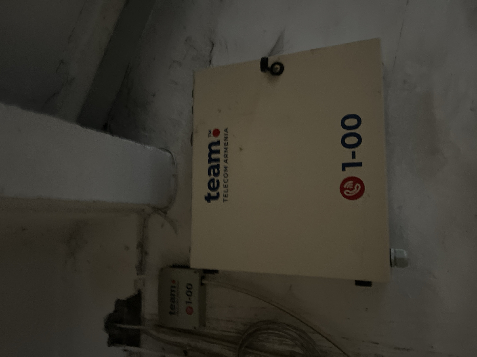 An indoor shot presents two white boxes mounted on a textured, light-colored wall in what appears to be Yerevan, Armenia. The larger, rectangular box bears the inscription "team TELECOM ARMENIA" and the phone number "01-00" vertically oriented on its right side. A smaller, similar box, is visible below the larger one. Various cables run out of these boxes. The wall shows signs of wear and disrepair with cracks. A concrete or masonry structure is visible on the left side of the frame.