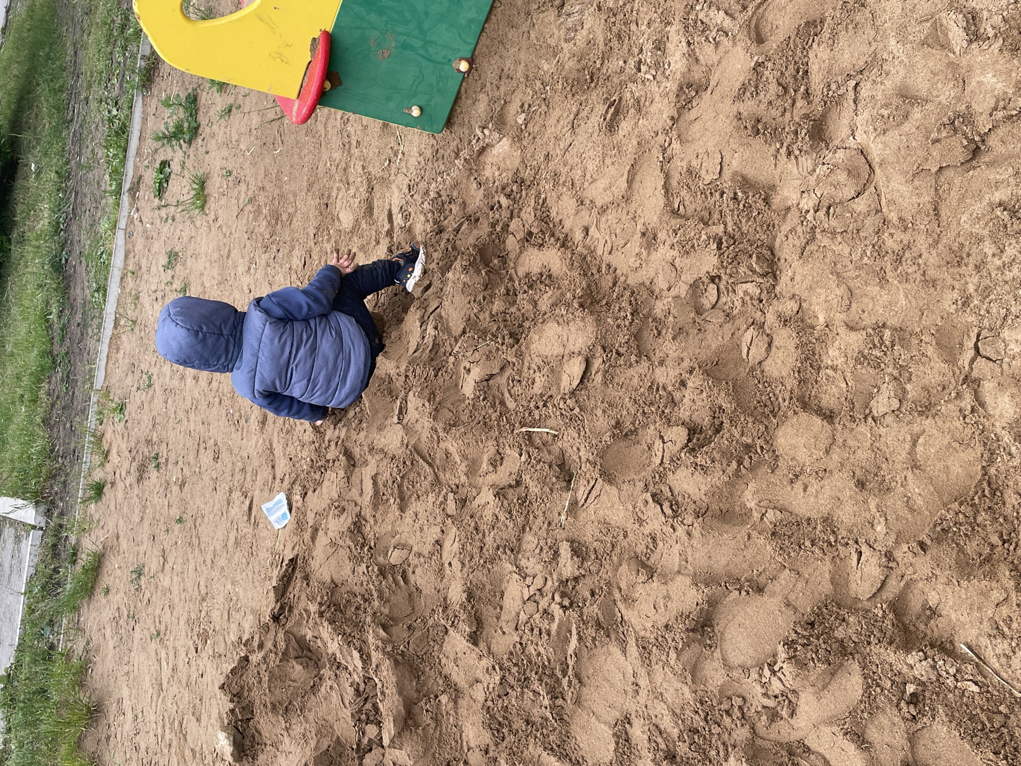 An overhead shot captures a child sitting or kneeling in a large sandy area, likely a sandbox, part of a playground in Perm, Russia. The child, viewed from behind, wears a blue hooded puffer jacket and dark pants, with their hands seemingly engaged with the sand. Their shoes, dark with some white details, are also visible.
The majority of the image is dominated by the light brown sand, which shows numerous footprints and impressions, indicating active play. The sand appears somewhat moist and clumpy in places. To the upper left, a portion of a colorful playground structure is visible, featuring bright yellow and green panels with red accents. Along the left edge of the sandbox, a strip of grass and dirt is present, bordered by a concrete curb or path, suggesting the edge of the play area. A small, crumpled white and blue object, possibly a wrapper or piece of paper, lies on the sand near the child.
The lighting suggests a daytime scene, possibly overcast, as there are no harsh shadows. No discernible text is visible in the image. The child's attire suggests cooler temperatures.