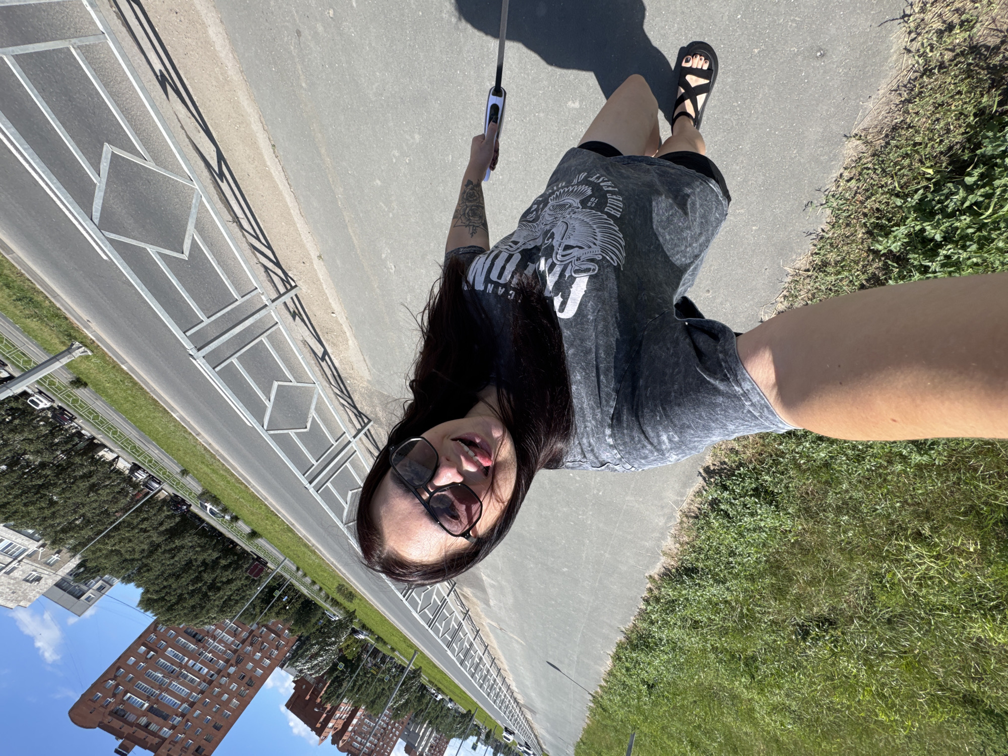 A young woman with dark hair, wearing sunglasses, a grey t-shirt, and black shorts, takes a selfie outdoors. She is holding a retractable dog leash in her right hand. The background shows a street in the city of Tolyatti, Russia, with buildings and trees visible. The woman appears to be walking on a sidewalk next to a road. The overall scene is a sunny day with a clear blue sky.
