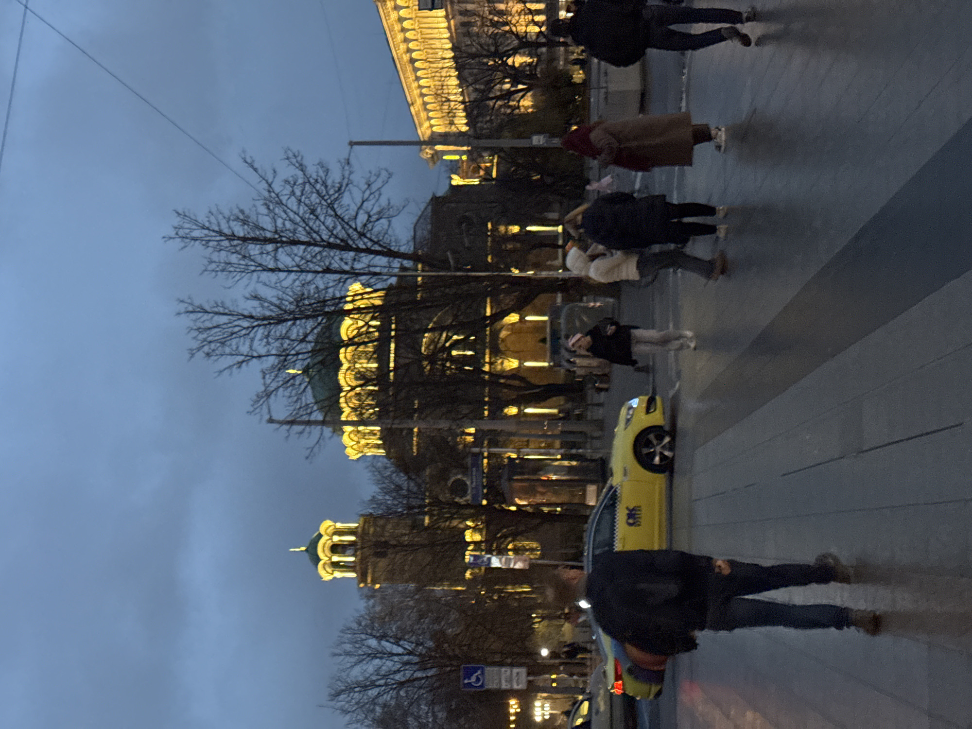 The image depicts a street scene in Sofia, Bulgaria, at dusk or early evening. The Alexander Nevsky Cathedral is prominently featured, illuminated and towering over the scene. Several pedestrians are walking on the paved sidewalk, some in groups, and others alone. A yellow taxi cab is parked along the street. The sky is overcast and grey. Bare trees are interspersed throughout the scene, adding a wintery feel. Streetlights and the illuminated cathedral provide the primary light sources.