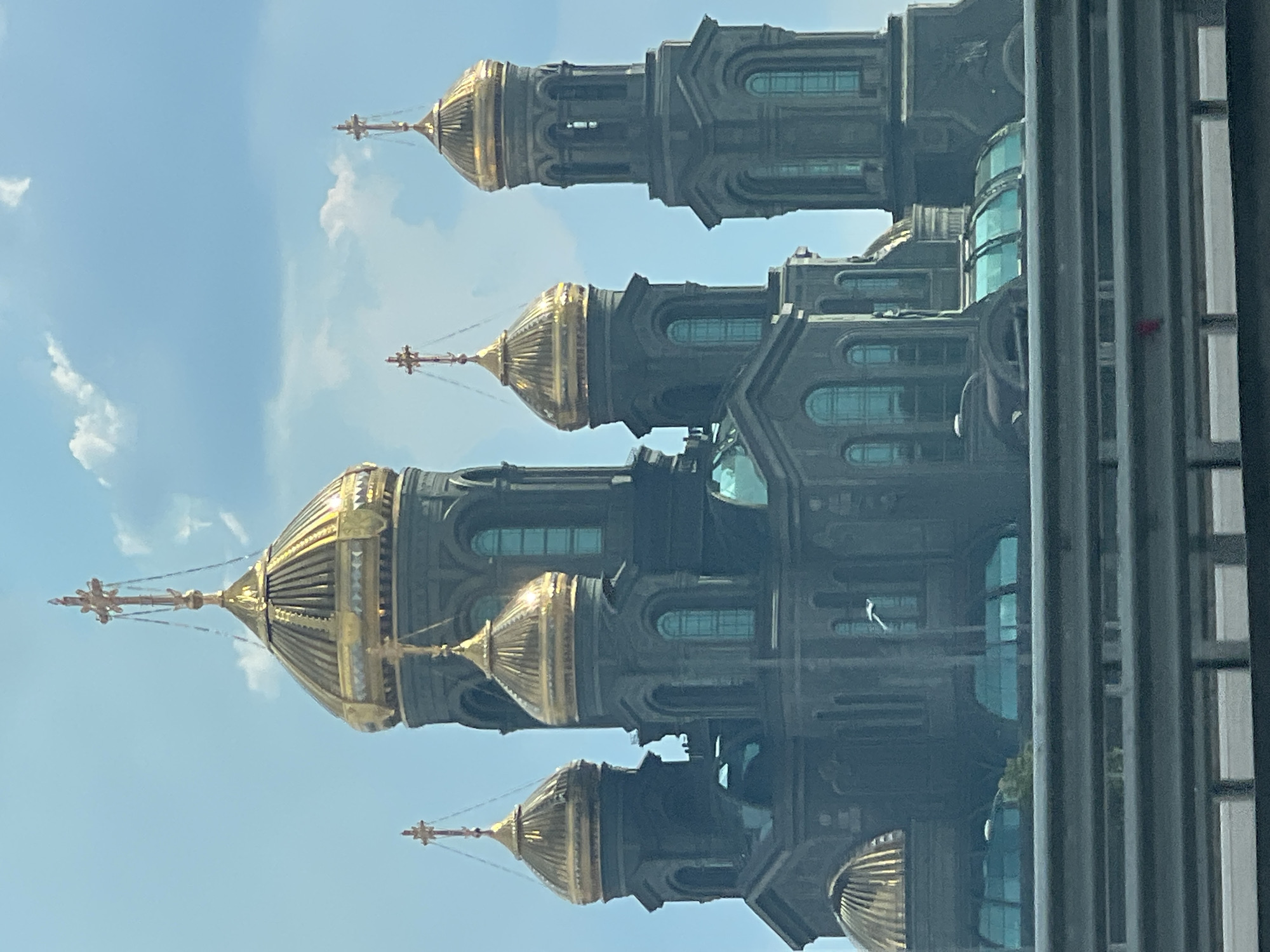 The image captures a view of the Main Cathedral of the Russian Armed Forces, located in Kubinka, Odintsovsky District, Moscow Oblast, Russia, though the prompt incorrectly states Zelenaya Roshcha, Russia. The cathedral is a grand, dark-colored structure with multiple golden domes and crosses. The sky above is a clear blue with scattered white clouds. In the foreground, a metallic guardrail runs horizontally across the frame, suggesting the photo was taken from a moving vehicle on a highway or elevated road. A bird is visible in flight in the middle distance, positioned between the cathedral and the guardrail. The architecture is ornate, with arched windows and intricate detailing on the towers and domes. The lighting suggests it is daytime, with sunlight glinting off the golden elements of the cathedral.
