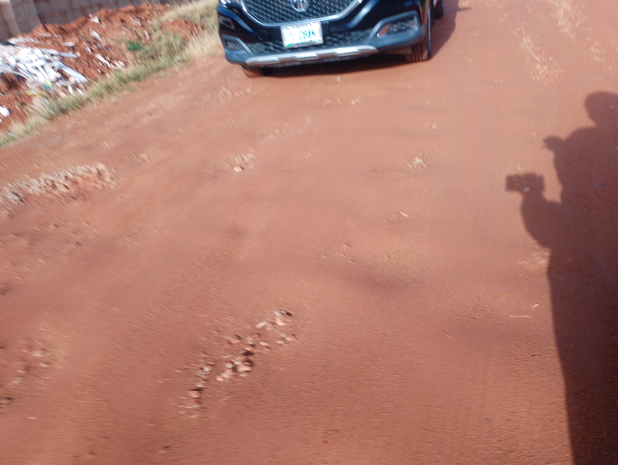 The image shows a close-up view of the front of a black SUV on a reddish-brown dirt road in Gura Top, Nigeria. The license plate of the vehicle is visible and reads "TRS 281N". In the background, to the left of the vehicle, is a pile of reddish-brown dirt mixed with debris, including white plastic items and some sparse green vegetation. To the right of the vehicle, a shadow of a person holding a phone is cast on the road. The road surface appears rough with small stones and indentations. The sky is not visible.