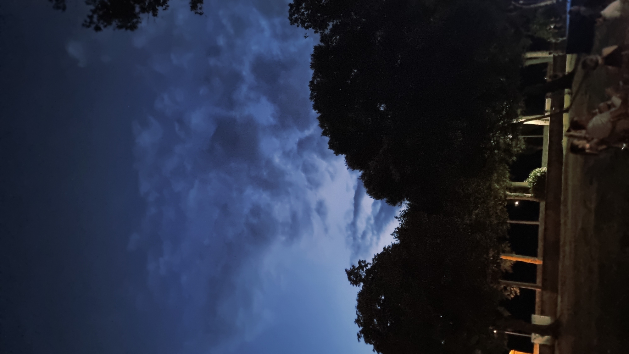 The image captures a dimly lit outdoor scene, likely in a park or public square, at dusk or night. The sky is a deep, moody blue with prominent, textured clouds, suggesting a dramatic or overcast evening. Silhouetted against the sky are the dark, lush canopies of several trees. In the foreground, a group of people are gathered, seated and interacting, though their features are indistinct due to the low light. The ground appears to be a grassy or paved area with some ambient lighting from unseen sources, casting subtle illumination. Behind the people and trees, a row of lamp posts or similar fixtures are visible, providing a faint, horizontal line of light. The overall atmosphere is serene and somewhat mysterious, with a focus on the interplay of light and shadow. The location is identified as Sant Joan Les Fonts, Spain.
