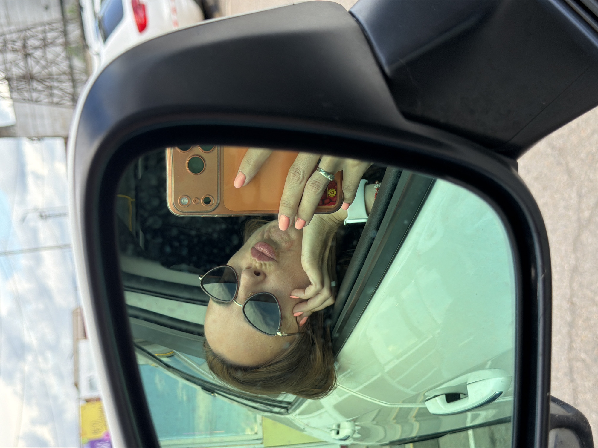 A woman takes a selfie in the side-view mirror of a car. She is wearing sunglasses and has pink nail polish on her fingernails. Her hair is brown and she has a ring on her finger. The background of the mirror shows a car and the sky. The photo appears to have been taken during the day. There is no visible text in the image.