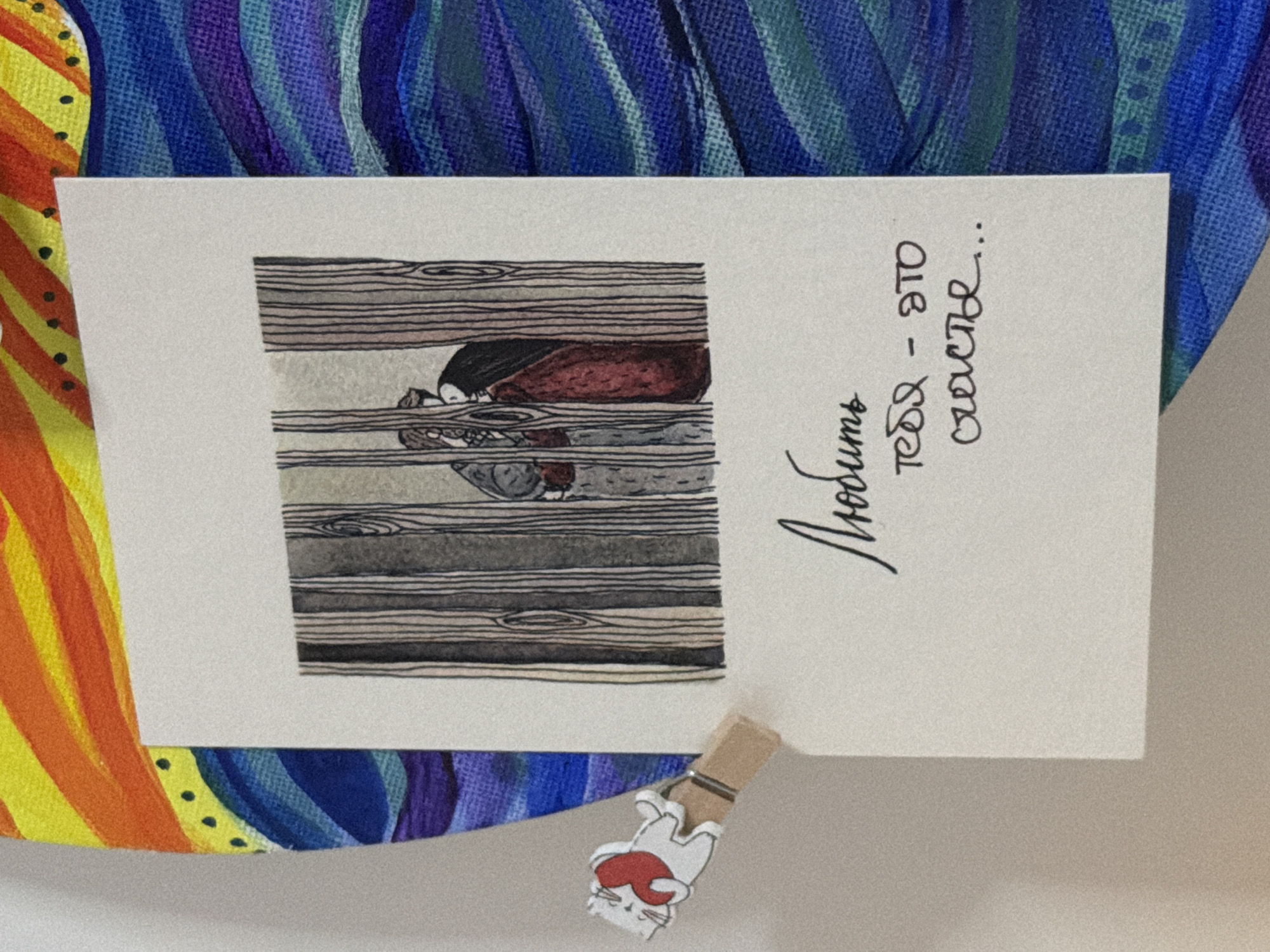 The image shows a white card with a drawing and handwritten text, clipped to a larger, colorful artwork by a wooden clothespin with a cat figure. The drawing on the card depicts two figures partially obscured by vertical wooden planks. The text on the card is in Russian and reads "Любить тебя - это счастье...", which translates to "Loving you is happiness...". The larger artwork features vibrant, abstract waves of color, with blues, purples, and yellows. The scene is indoors, with a plain white wall visible to the left. There is no discernible location in Moscow, Russia.