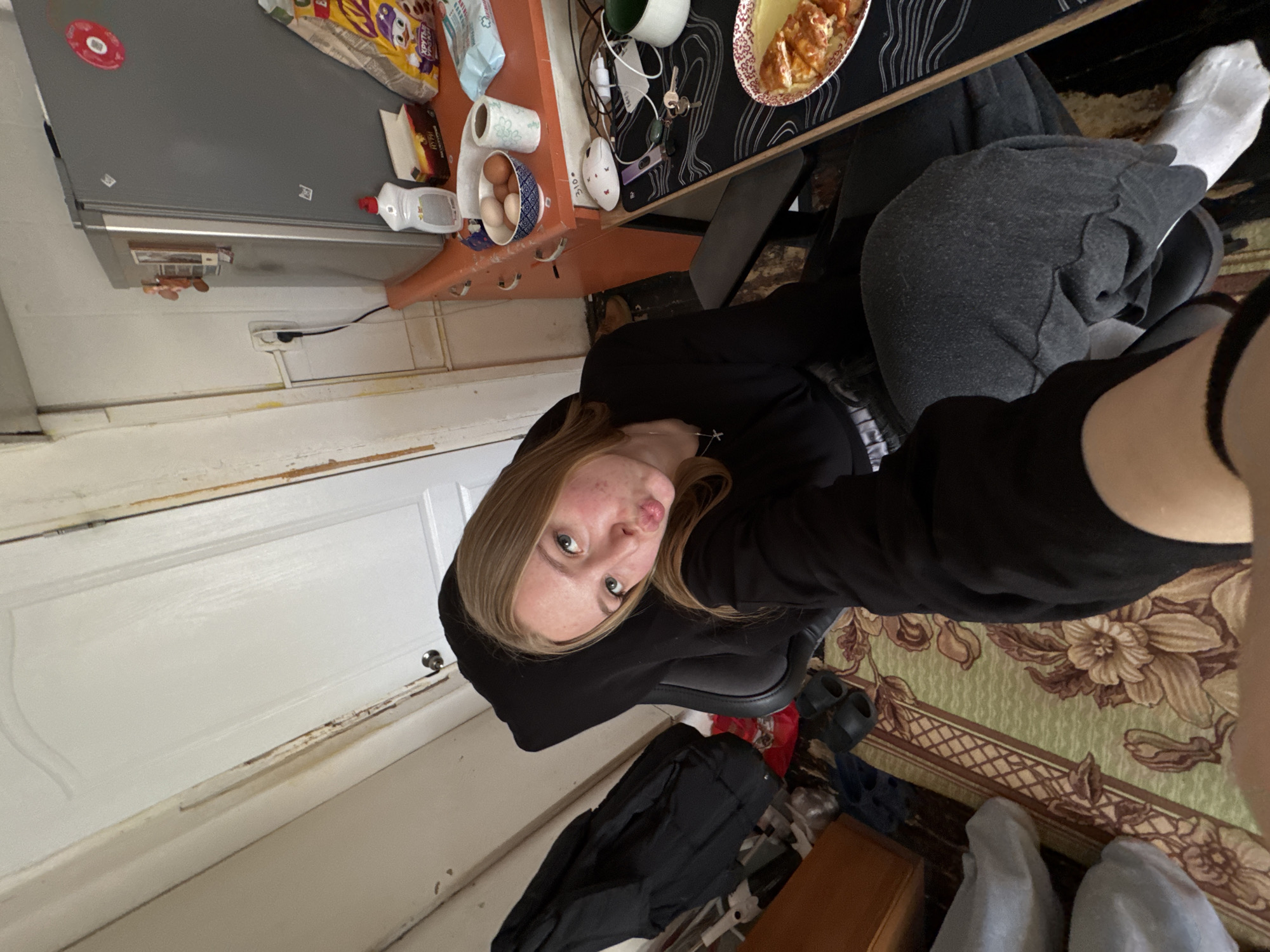 A young woman is taking a selfie indoors, likely in an apartment in Perm, Russia. She is sitting in a black office chair, wearing a black hooded sweatshirt and gray sweatpants. Her legs are crossed, and she is wearing white socks. A wooden desk is positioned in front of her, with a plate of food, a computer mouse, keys, and other miscellaneous items. The desk has a black surface with a pattern. Behind the desk and the woman is a rug with a floral pattern. The room is cluttered with various objects including a black bag on the floor, shoes, and a wooden cabinet. A white door is visible on the left side of the image, and a refrigerator is located in the upper left corner. The general impression is a casually untidy domestic scene.