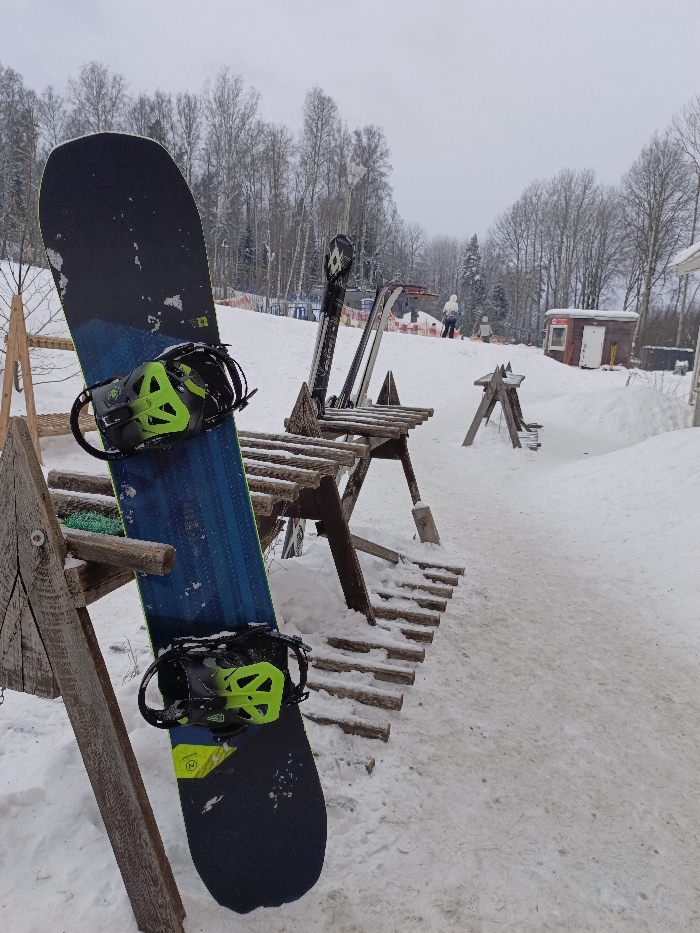 The image portrays a winter scene at a ski resort or recreational slope in Svetloe, Russia, under an overcast sky. In the immediate foreground, a dark blue and turquoise snowboard, equipped with neon green and black bindings, leans against a rustic wooden ski rack positioned on the left. To its right, another similar wooden rack holds at least two pairs of skis upright, one pair distinctly black and white. The ground is primarily covered in packed, granular snow, with some areas showing exposed patches of dirt or underlying ground.
The midground features an uphill snow-covered slope. Two individuals are discernible ascending the slope in the distance; one person is dressed in white outerwear, and a second, smaller figure is positioned slightly behind and to the right of the first. On the right side of the slope, a small, reddish-brown wooden utility building with a white door and a red emblem above it is visible. An orange safety net extends along the left perimeter of the slope, bordering a line of bare deciduous trees. A dense forest of bare trees forms the distant background, blending with the pale grey sky.