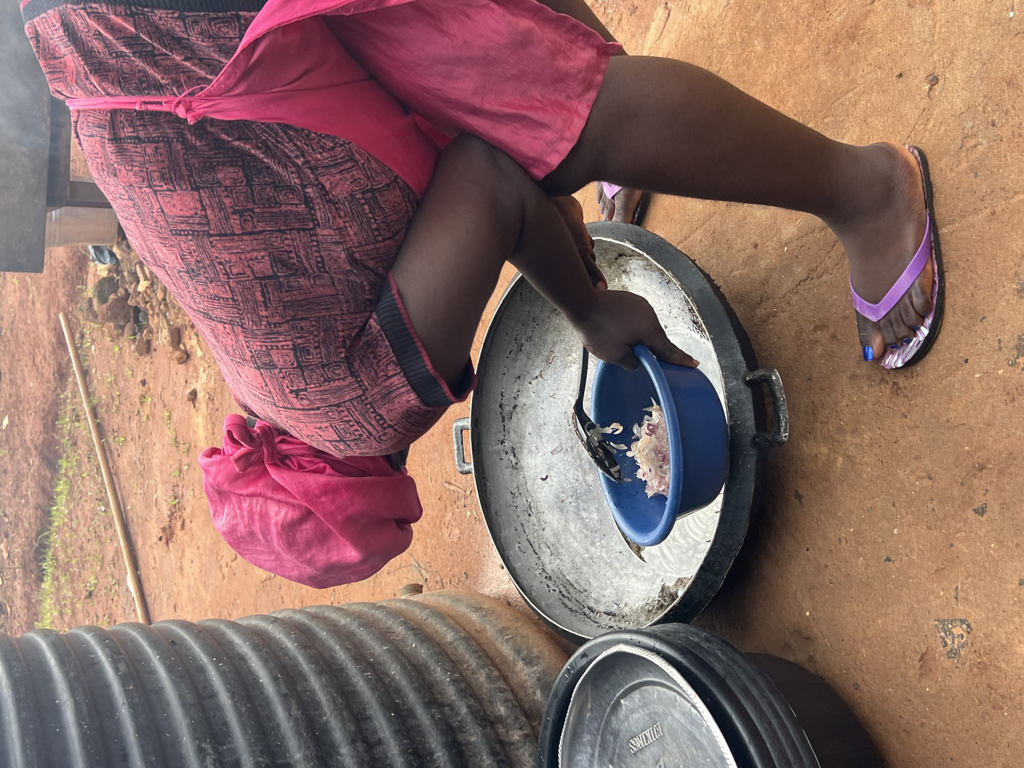 A woman wearing a pink headscarf, a patterned pink and black dress, and a pink apron leans over a large metal pot in Nkpor, Nigeria. She is holding a blue bowl containing chopped onions with a black spatula, preparing to add them to the pot. Next to the pot are stacked black plastic containers and lids. The ground is made of reddish-brown dirt. In the background, a large black cylindrical water tank is visible, along with some vegetation and rocks. The woman is wearing purple flip-flops.