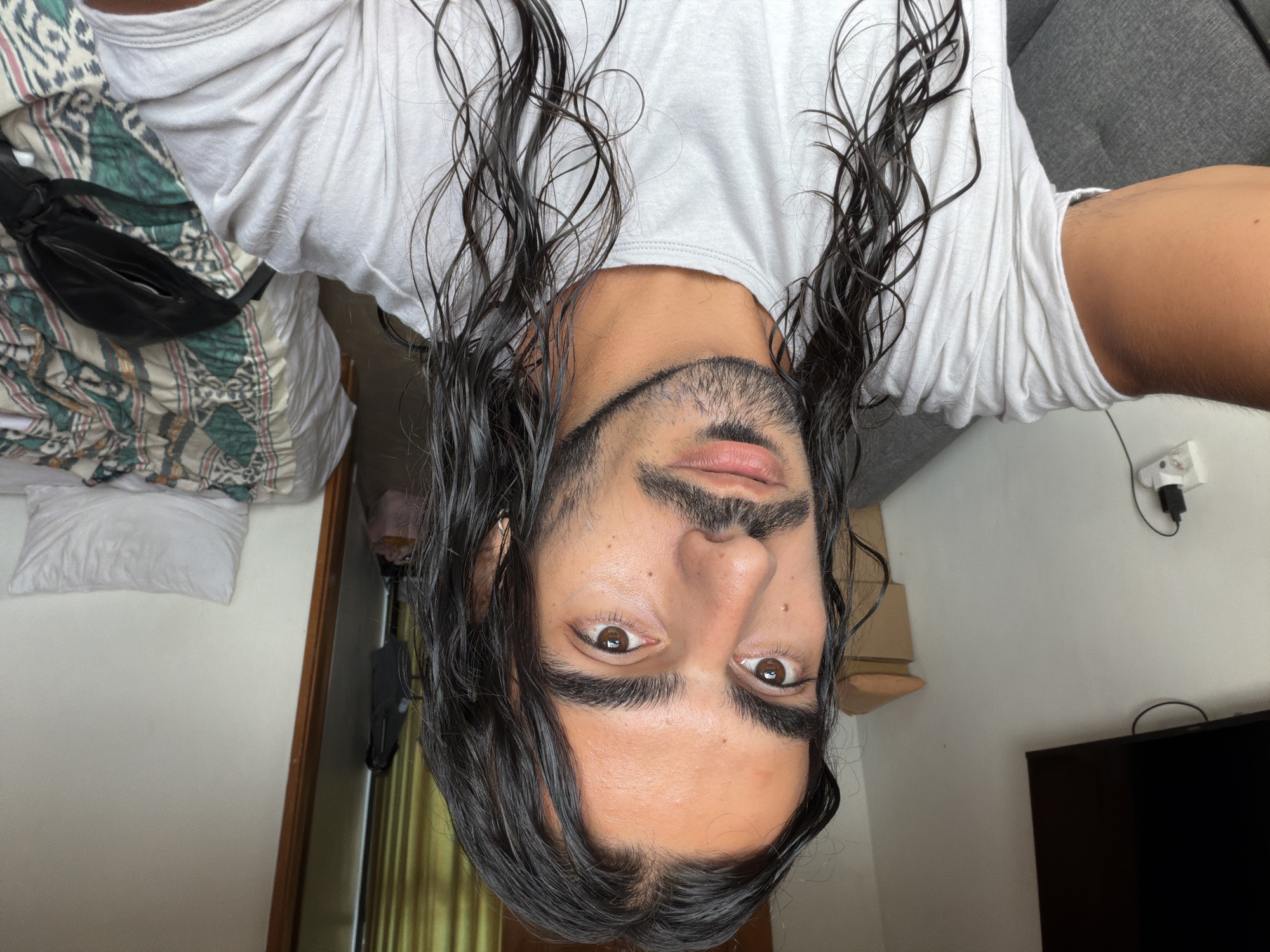 A selfie of a man with long, wet, dark hair and a beard, taken from an inverted angle. He is wearing a white t-shirt. The background reveals a bedroom interior. To the left, there's a bed with a pillow and a patterned blanket, along with a black fanny pack. To the right, there's a gray sofa, a wall with a power adapter, a cardboard box on a shelf, and the corner of a black TV. The room is in North Kuta, Indonesia.