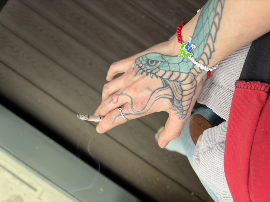 The image depicts a close-up, high-angle view of a person's left hand and lower body, situated in Sofia, Bulgaria. The left hand holds a lit cigarette between the thumb and index finger, from which a visible plume of smoke emanates. The cigarette features a prominent column of ash. A silver-toned ring with a small red stone is worn on the ring finger, and a red heart outline tattoo is visible on the index finger. The back of the hand and forearm are extensively tattooed with a stylized dragon or serpentine design, colored light blue with black outlines. Two braided bracelets, one red and one white, adorn the wrist. A green, purple, and black 'The Hulk' charm is affixed to the red bracelet. The subject is wearing a red garment on the torso, visible with a black hem, and light grey corduroy trousers that feature a brown oval patch on the inner thigh. A partial view of light-colored footwear is discernible at the lower left. The background consists of a dark, horizontally planked surface, consistent with a wooden deck or similar flooring, and a blurred, dark-framed glass or reflective surface in the upper left quadrant.