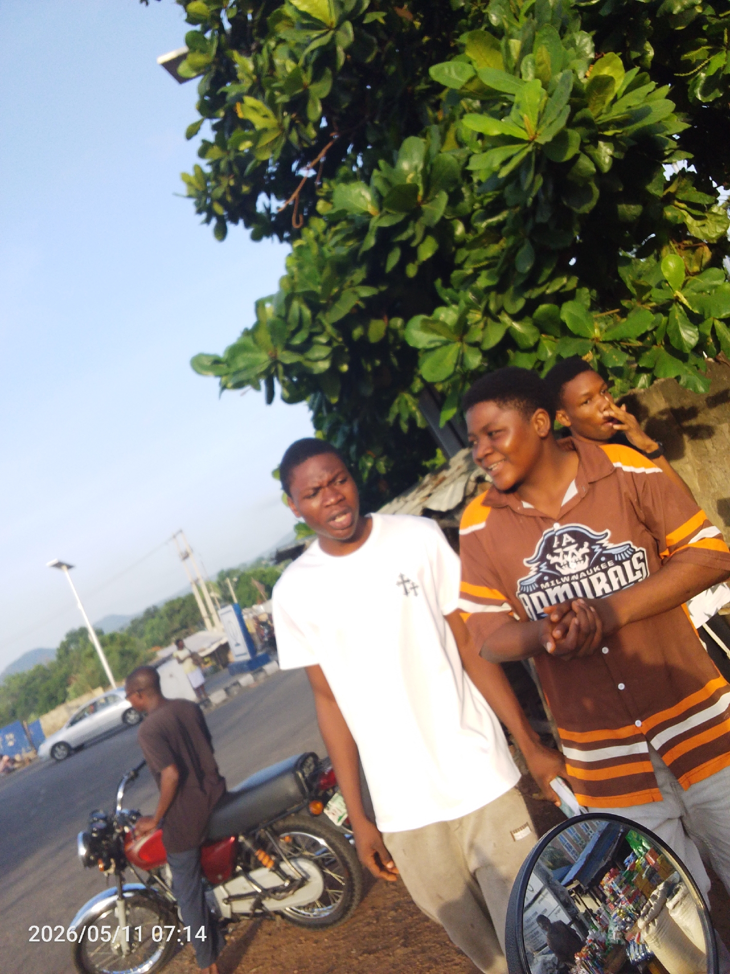 The photo depicts four young men in an outdoor setting in Adankolo, Nigeria. One man is seated on a parked red motorcycle, wearing a dark shirt and pants. In the foreground, two men are standing; one is wearing a white t-shirt with a symbol on the chest, and the other is wearing a brown button-down shirt with orange stripes and a "Milwaukee Admirals" logo. A fourth man is visible behind them near a wall. A lush green tree occupies the upper right corner of the frame, partially obscuring a building. A street, a car, and buildings are visible in the background. The date "2026/05/11 07:14" is printed on the bottom left of the image.
