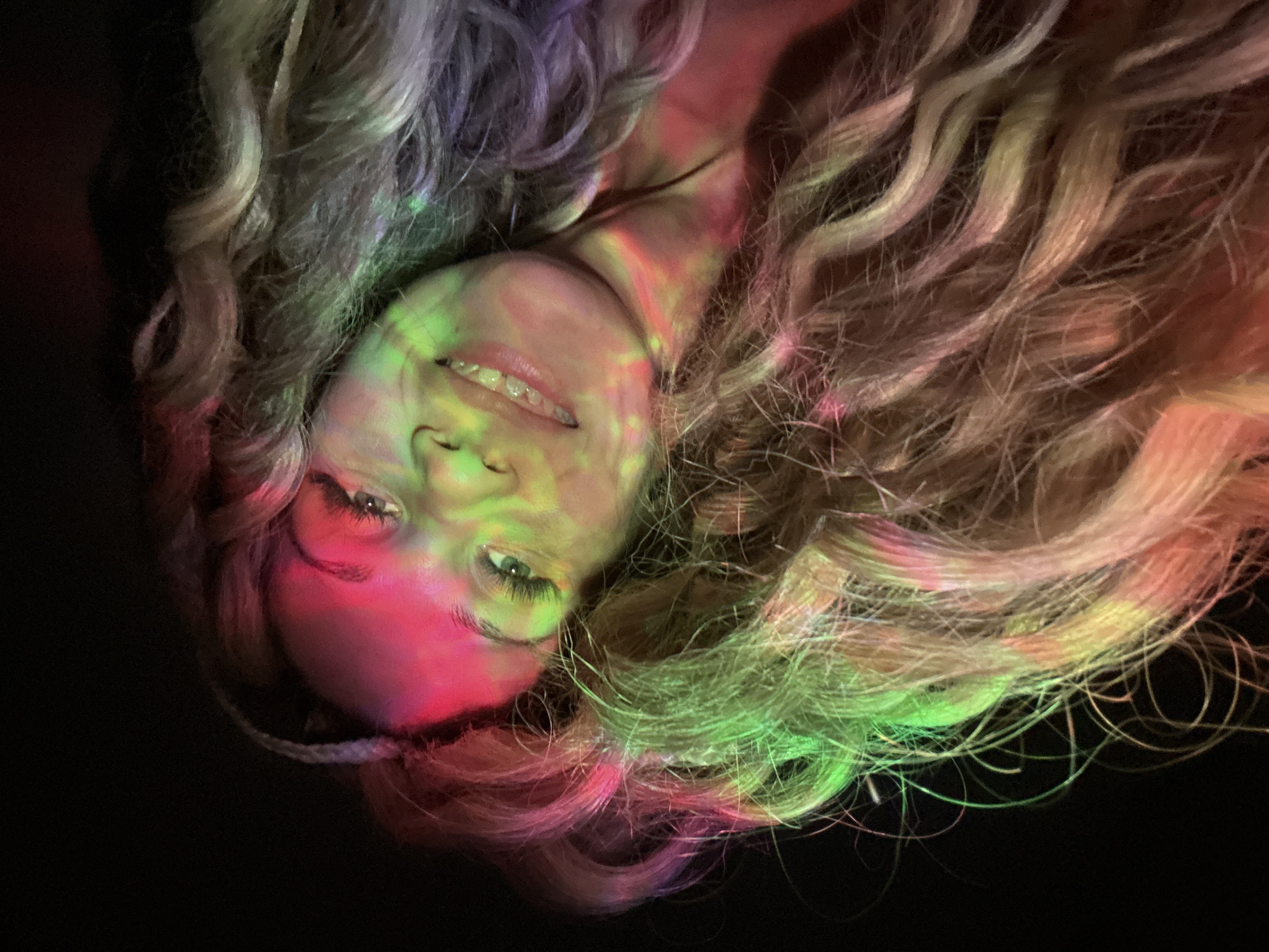 A woman with wavy blonde hair lies on a dark surface, possibly a floor or table, with her head angled towards the viewer. Her face is illuminated with green and pink light, giving a neon-like effect. Her eyes are open, and she has a faint smile. The hair is spread around her head, with strands extending into the dark background. A light-colored cord or decorative braid is visible near the bottom of the frame, close to her head. The photograph is taken at a close range. The city of Rusanovo, Russia is not discernible from the image.