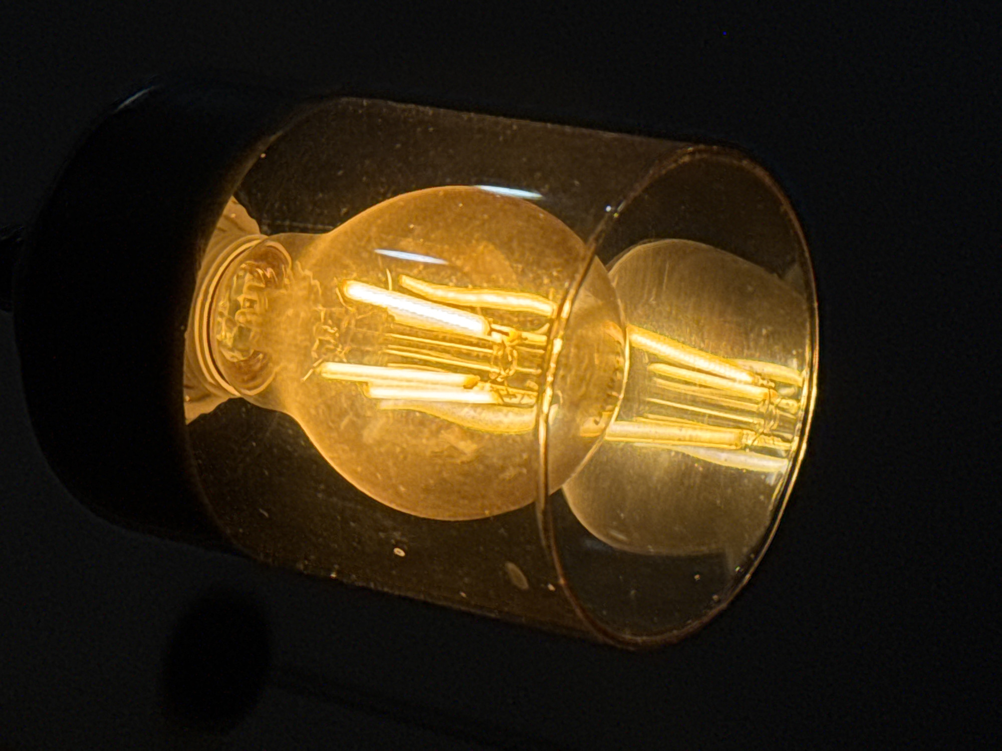 The image shows a lit vintage-style light bulb encased in a clear glass cylindrical shade. The bulb is attached to a dark, possibly black, fixture. The bulb's filaments are visible and glowing with a warm, yellow light. The background is dark, emphasizing the brightness of the bulb. The image does not contain information about the location of Novo-Nikol'skoe, Russia, and this cannot be inferred from the visible content.