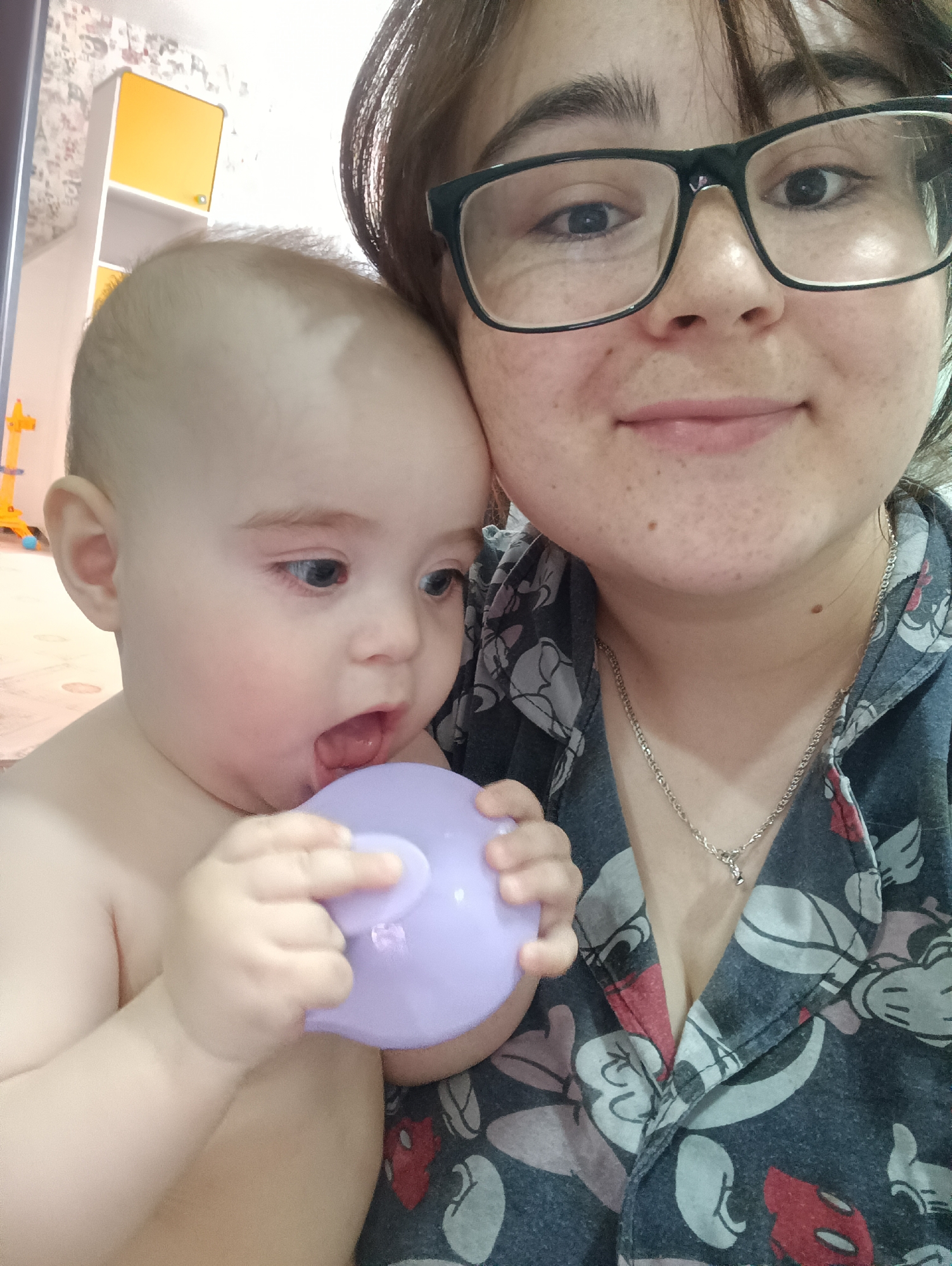 A close-up shot features a woman with dark-framed glasses holding a baby. The baby, who appears to be teething, is chewing on a lavender-colored object. The woman is wearing a button-up shirt patterned with cartoon characters. A silver necklace is visible around her neck. The background shows a room with a yellow and white cabinet. The location is Unknown, Unknown.