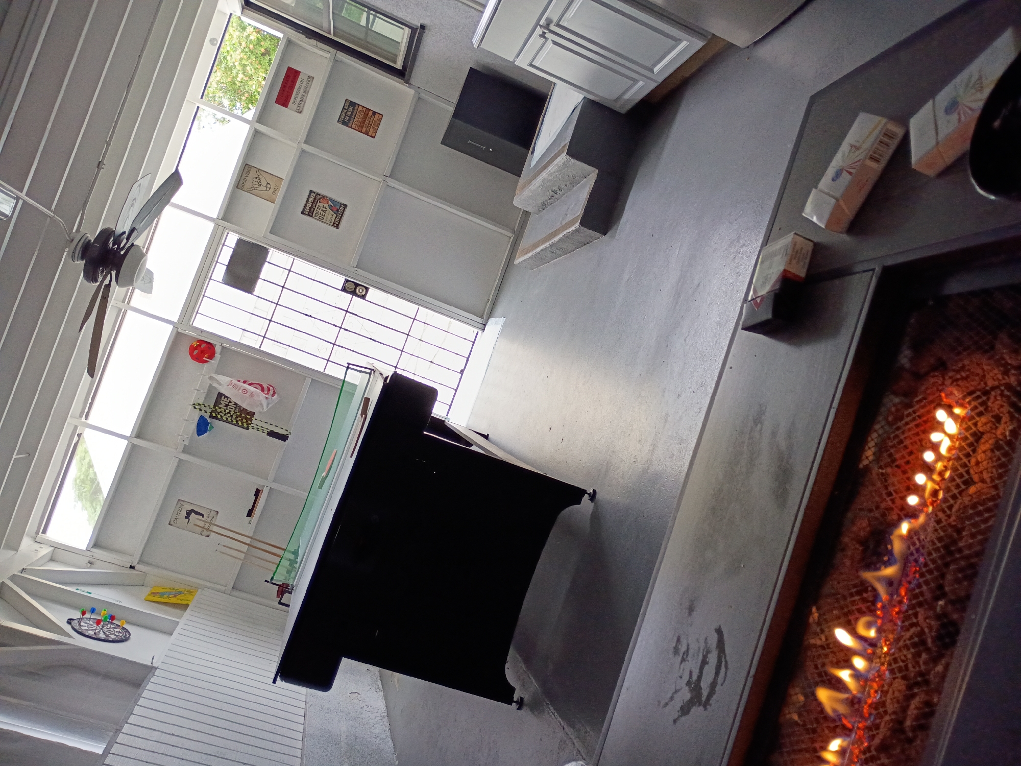 Here is a forensic description of the image:
The image depicts an indoor game room or entertainment space, with a variety of recreational elements. In the foreground to the right is a linear fireplace with visible flames behind a screen or grate. Several packs of cigarettes or similar small rectangular packages sit on the surface next to the fireplace. Adjacent to the fireplace is a dark-colored pool table. Above the pool table, various items and decor are mounted on the white-painted walls including a dartboard, pool cues, a promotional poster, and other decorative items. The upper area of the room shows a series of framed signs and artwork. There is a ceiling fan in the upper-left corner. The room seems to have an abundance of natural light coming in through windows near the ceiling.
It is not possible to determine the precise location within the city of Los Angeles, United States of America, solely from the given information.