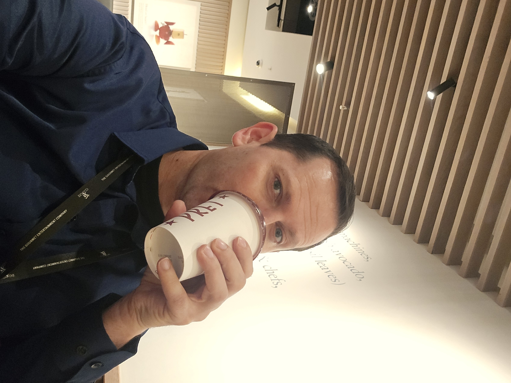 A man is drinking from a white paper cup with "PRET" printed on it in burgundy. He is wearing a dark blue button-down shirt and a lanyard with the words "THE GOURMET ENTERTAINMENT COMPANY" and "DONCO" printed on it. The background features a light beige wooden slat wall with modern black track lighting. A white wall has text printed on it, including "machines, avocado, leaves" and "chefs". The scene is most likely set in London, United Kingdom, as PRET A MANGER is a common coffee shop chain in the city. The photograph is taken at an angle, with the man and the wall slightly tilted.