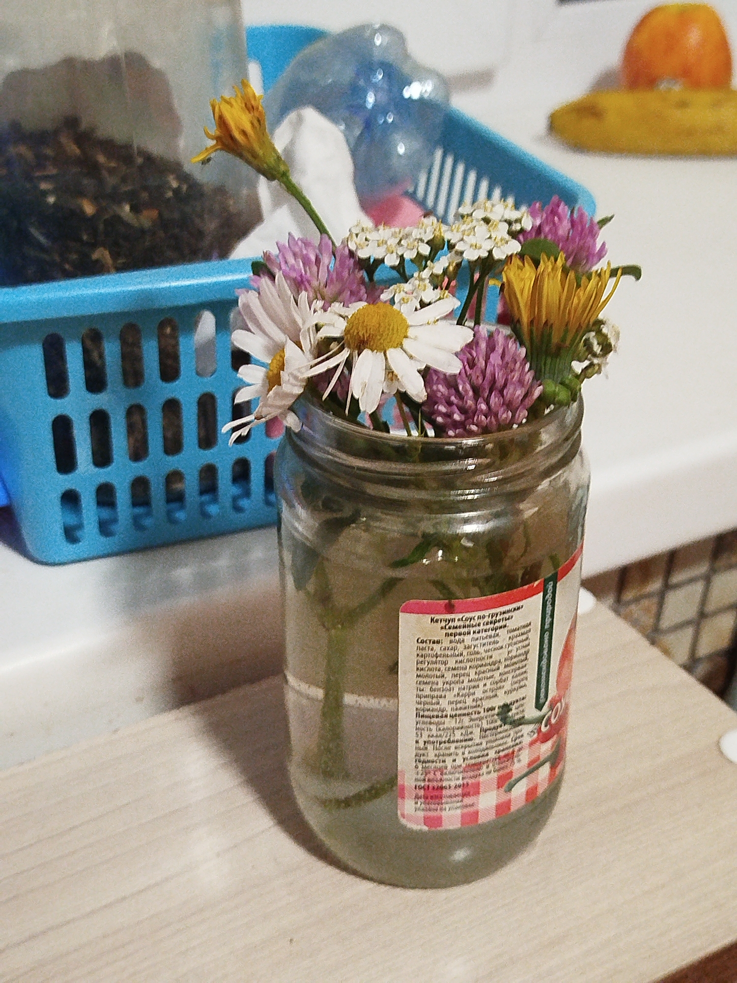 In a domestic setting, possibly a kitchen, a clear glass jar holds a small bouquet of wildflowers, including daisies, clover, and yellow dandelions. The jar, a repurposed food container, displays a label with Russian text, partially obscured by the flowers. Behind the jar, a bright blue plastic basket contains dried organic material, possibly tea leaves or herbs. In the background, a blurry orange fruit, likely a kumquat or small orange, rests on a white surface. The overall scene is dimly lit, with soft shadows cast across a light-colored wooden countertop.