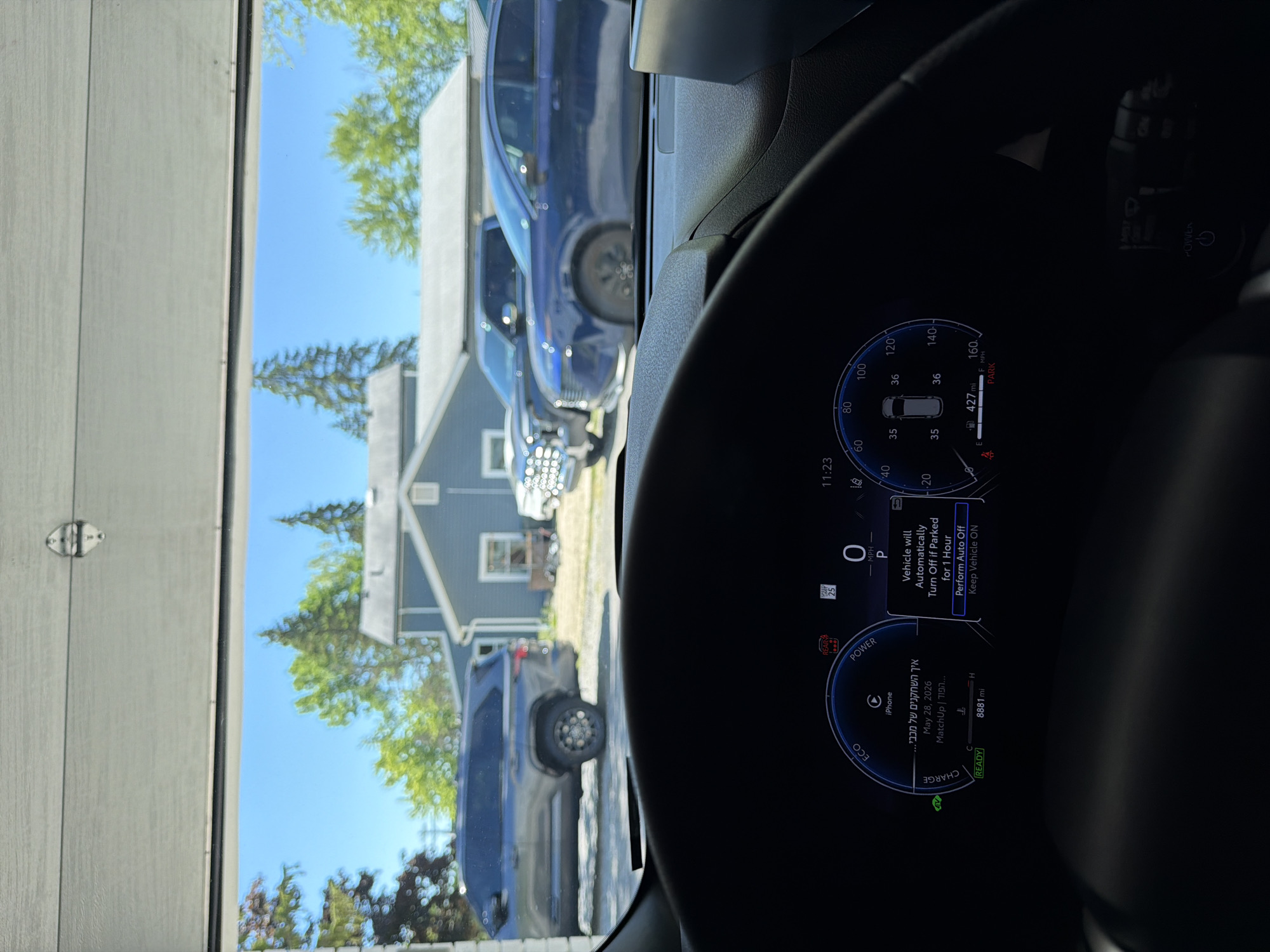The image captures a first-person perspective from the driver's seat of a parked vehicle, looking out through an open garage door in Pepper Pike, United States of America. The time displayed on the vehicle's digital dashboard is 11:23. The dashboard prominently shows "8881 mi" on the odometer, with the vehicle in "P" (Parked) mode and indicating "O MPH". An "EV" mode is active, and a "READY" status is displayed. A notification bubble states "Vehicle will Automatically Turn Off if Parked for 1 Hour," offering options to "Perform Auto Off" or "Keep Vehicle ON." Other dashboard indicators include a low fuel warning, a remaining range of "427 mi," and a "REAR & SIDE" warning light. A temperature reading of "25" is visible, along with text "iPhone" and Hebrew text "איך השחקנים של מכבי..." (How are the players of Maccabi...), a date of "May 28, 2026," and "MatchUp | הפוך..." (MatchUp | Reverse...).
Outside the garage, under a clear blue sky, three vehicles are parked on a paved surface. To the left, a dark gray large SUV (possibly a Toyota Sequoia) is visible. To its right, a white pickup truck is parked. Further to the right, a dark blue sedan is positioned. In the background, a light blue house with a white roof is visible behind the parked vehicles. Beyond the structures, various green trees, including both coniferous and deciduous types, indicate a vibrant summer or late spring environment. The lower section of a light-colored garage door is visible at the top of the frame, with a metal hinge mechanism present on the top right.