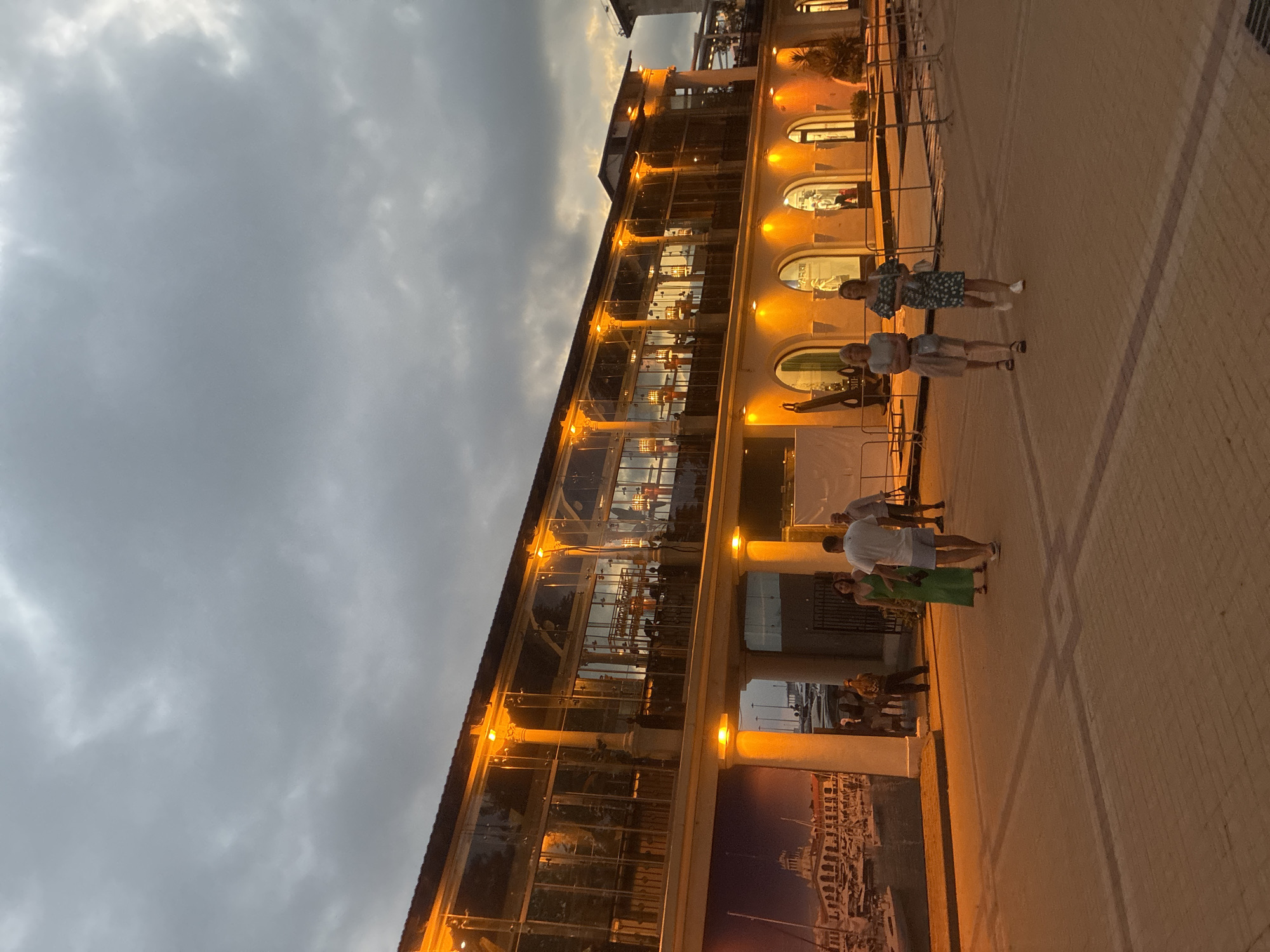 The image depicts a street scene in Sochi, Russia, at dusk. A modern building with large glass windows and warm lighting stands prominently on the right side of the frame. People are walking on a brick-paved walkway in front of the building. The sky is cloudy and overcast, typical of the evening weather. The building appears to be a commercial establishment, possibly a restaurant or shops, with visible interior lighting and furnishings. The overall atmosphere is calm and casual, with individuals engaged in leisurely strolls.