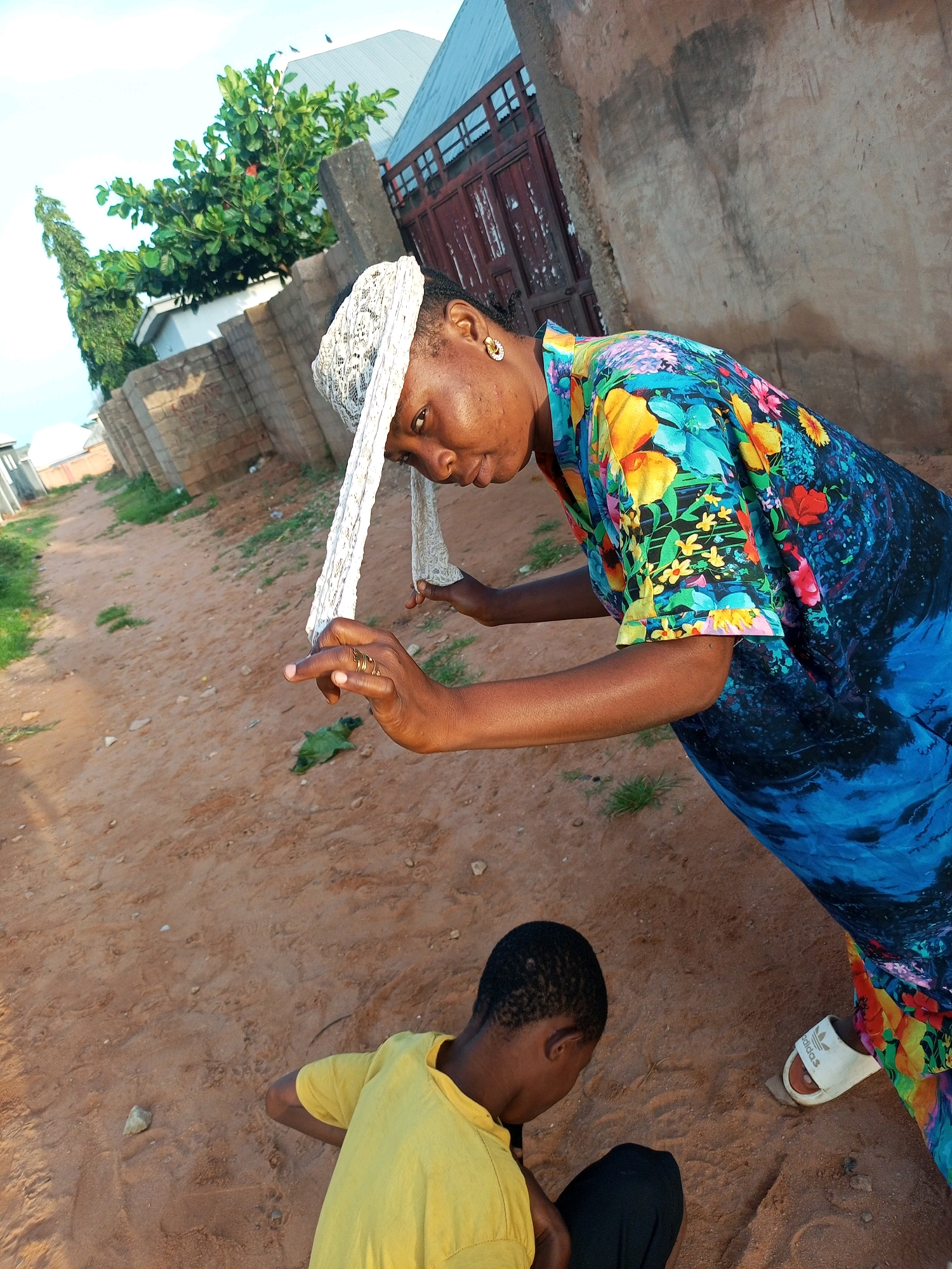 A young African child wearing a yellow t-shirt and black pants is crouching on the sandy ground. Beside him, a woman with a white head covering and a colorful blue, green, and floral dress is leaning over, looking down at the child. Her left hand is extended and she is wearing rings on her fingers. The scene appears to be outdoors in a dusty or sandy area, possibly a street or yard, with a brick wall and a building in the background. The overall lighting suggests it is daytime. The location is identified as Tempure, Nigeria.