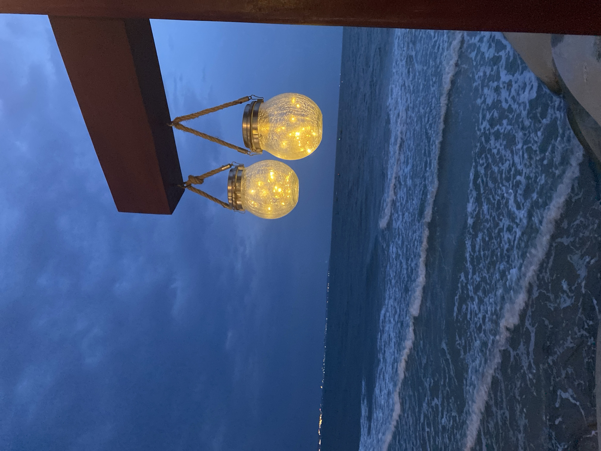Two illuminated, glass orb lanterns hang from a brown wooden beam. The background features a stormy, dark blue sky and an ocean with white-capped waves. The scene appears to be captured from a pier or balcony. The lights of Phu Thuy Ward, Vietnam, are visible in the distance along the shoreline.