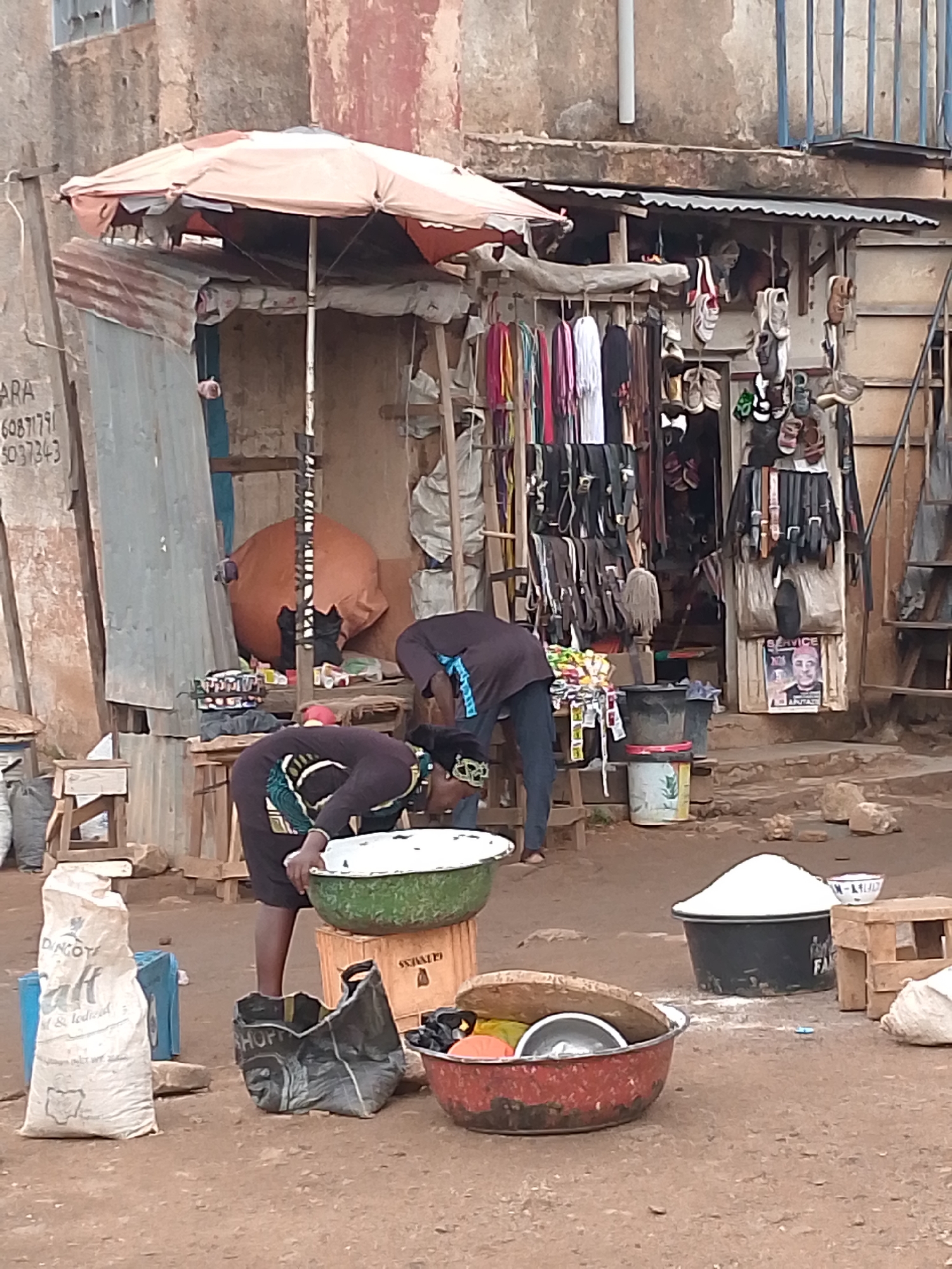 The image depicts an outdoor market scene in Bukuru, Nigeria. Two people are positioned near a green enamel bowl on top of a Guinness crate. A red enamel bowl, various bags (including one labeled "SHOP"), and a bag of "DINGOTE Salt" are on the ground. To the right, a dark grey container is piled with what looks like white powder, possibly sugar or salt, and a small wooden stool is nearby.
In the background, a makeshift shop is visible. It has a tattered orange umbrella providing shade. Scarves, belts, and sandals are displayed. The facade of the shop includes a poster with a portrait.
The general environment appears urban with buildings. The ground is a dirt surface.