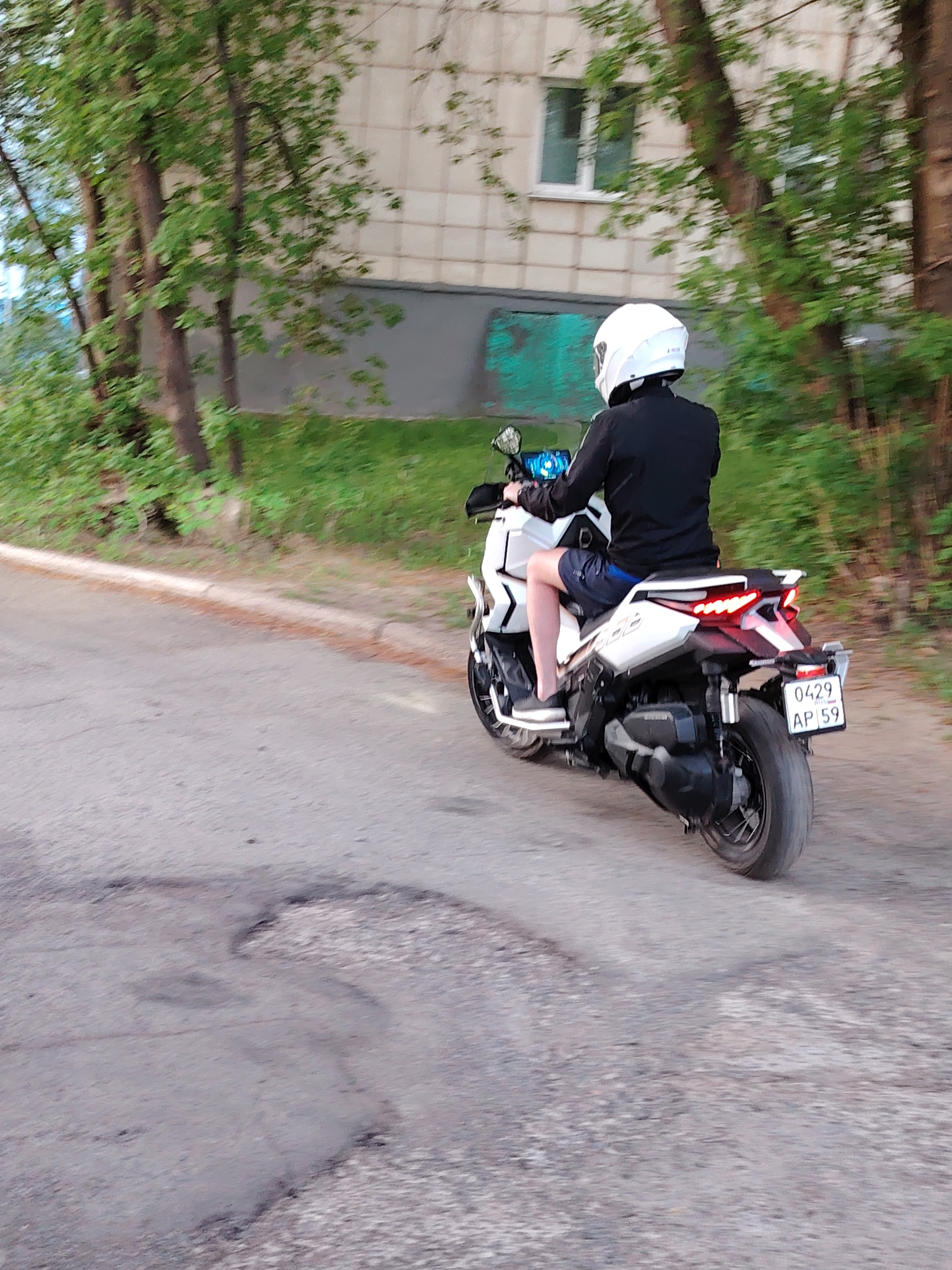 The image depicts a person riding a white scooter on a paved road. The rider wears a white helmet and dark clothing. The scooter has a visible license plate number, "0429 AP 59." The road surface shows signs of wear and damage. Trees and a building are visible in the background. Based on the license plate region code "59," and the greenery, the location is Chaikovsky, Russia.