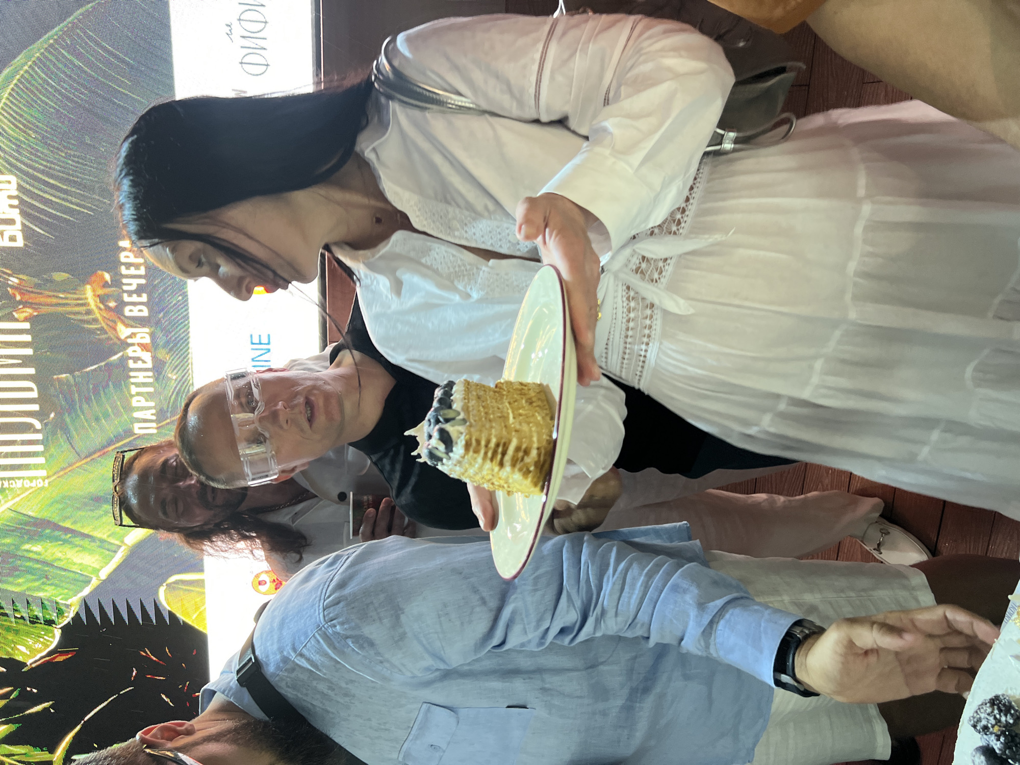 The image shows a group of people gathered, possibly at an event, with a screen displaying text and graphics in the background. A woman in a white dress holds a plate with a slice of layered cake topped with blueberries. To her left, a man in a light blue shirt and white shorts is visible, and to her right, another person is partially obscured. In the background, the screen appears to display event details or advertisements, with some text readable in Russian, potentially indicating a location in Sochi, Russia. The overall scene suggests a social gathering or celebration.