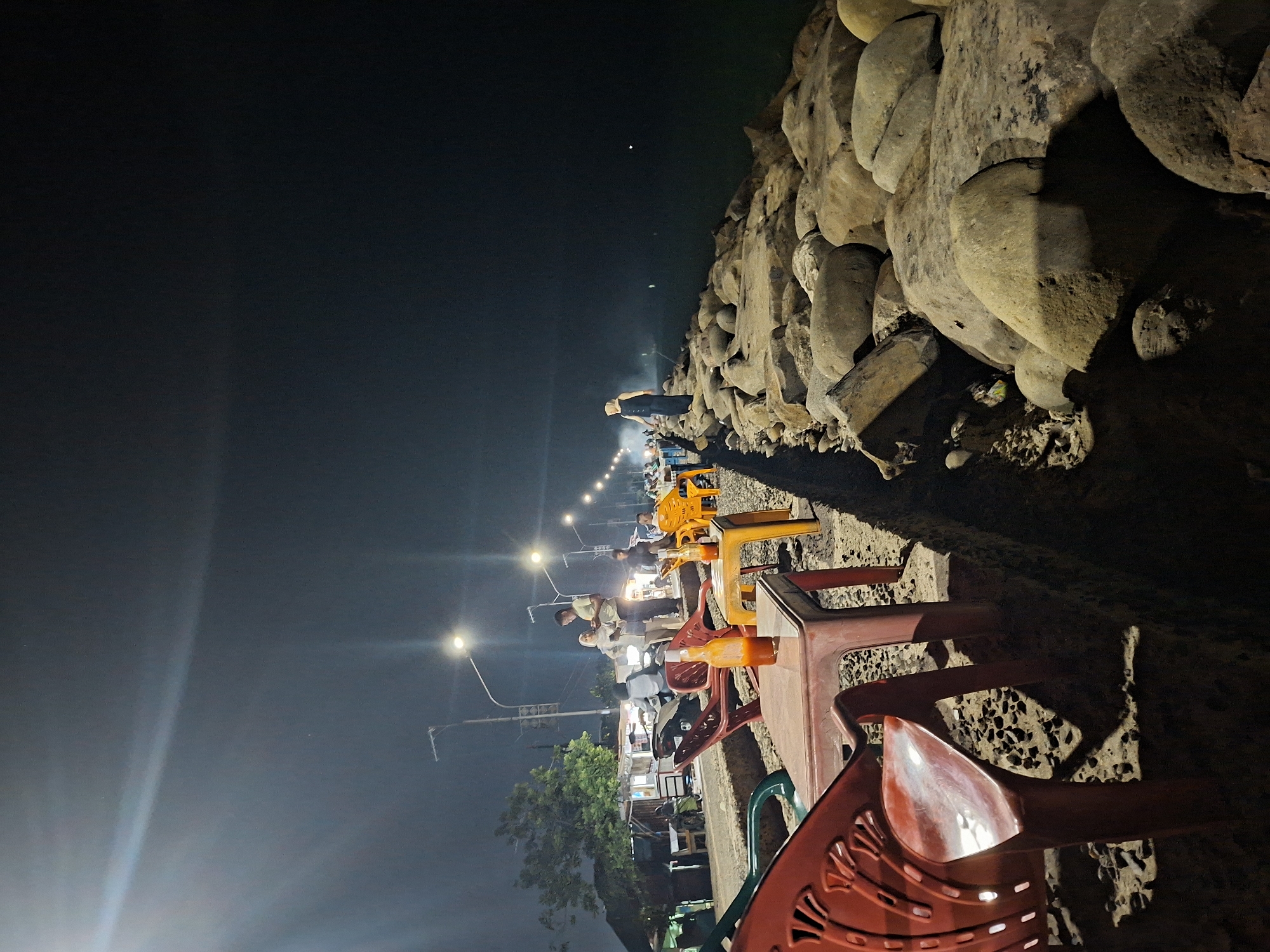 The image depicts a nighttime scene at a cafe or eatery near the coast in Lhokseumawe, Indonesia. The foreground contains a row of brightly colored plastic chairs and tables, primarily orange and red. These chairs seem to be arranged for patrons to sit and enjoy the view. In the background, a row of streetlights illuminates the area, highlighting figures that appear to be people standing or sitting along the waterfront. Along the right side of the frame is a large, irregular wall of large rocks or boulders, bordering the waterfront. The overall atmosphere suggests a casual, outdoor dining experience by the sea in Lhokseumawe. The night sky is dark, with a few visible stars, and the water extends into the darkness in the distance.