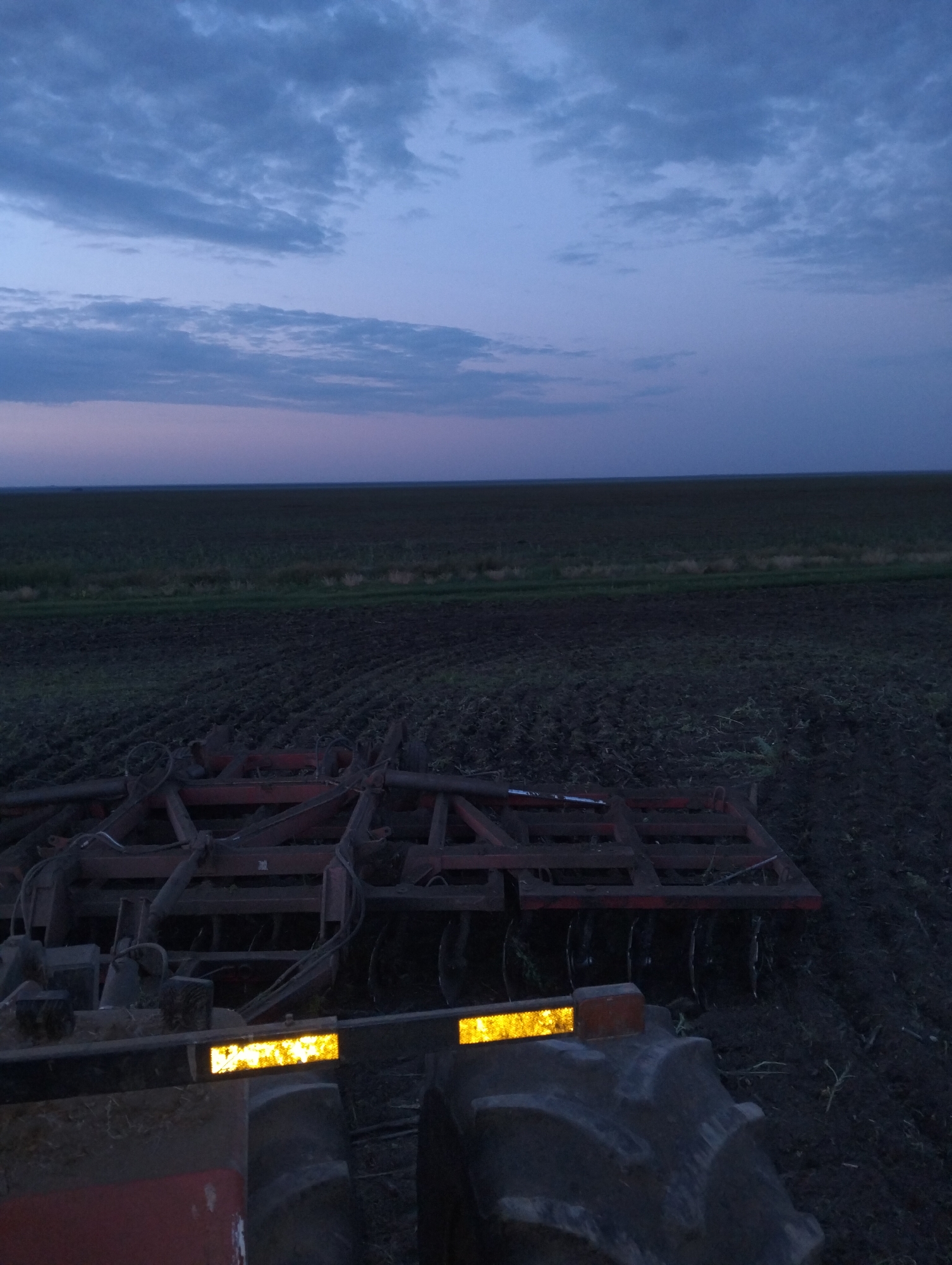 An outdoor, low-angle, full shot captures a farm field at dusk. The sky is a deep blue with scattered clouds, transitioning to a faint pinkish hue near the horizon. In the foreground, a portion of a red farm tractor is visible, with a large tire and part of the implement attached to it. The implement appears to be a disc harrow, with multiple discs visible. The field itself is dark soil, with rows of tilled earth. A dark, horizontal line in the distance suggests a treeline or further fields. The lighting indicates it is twilight.
