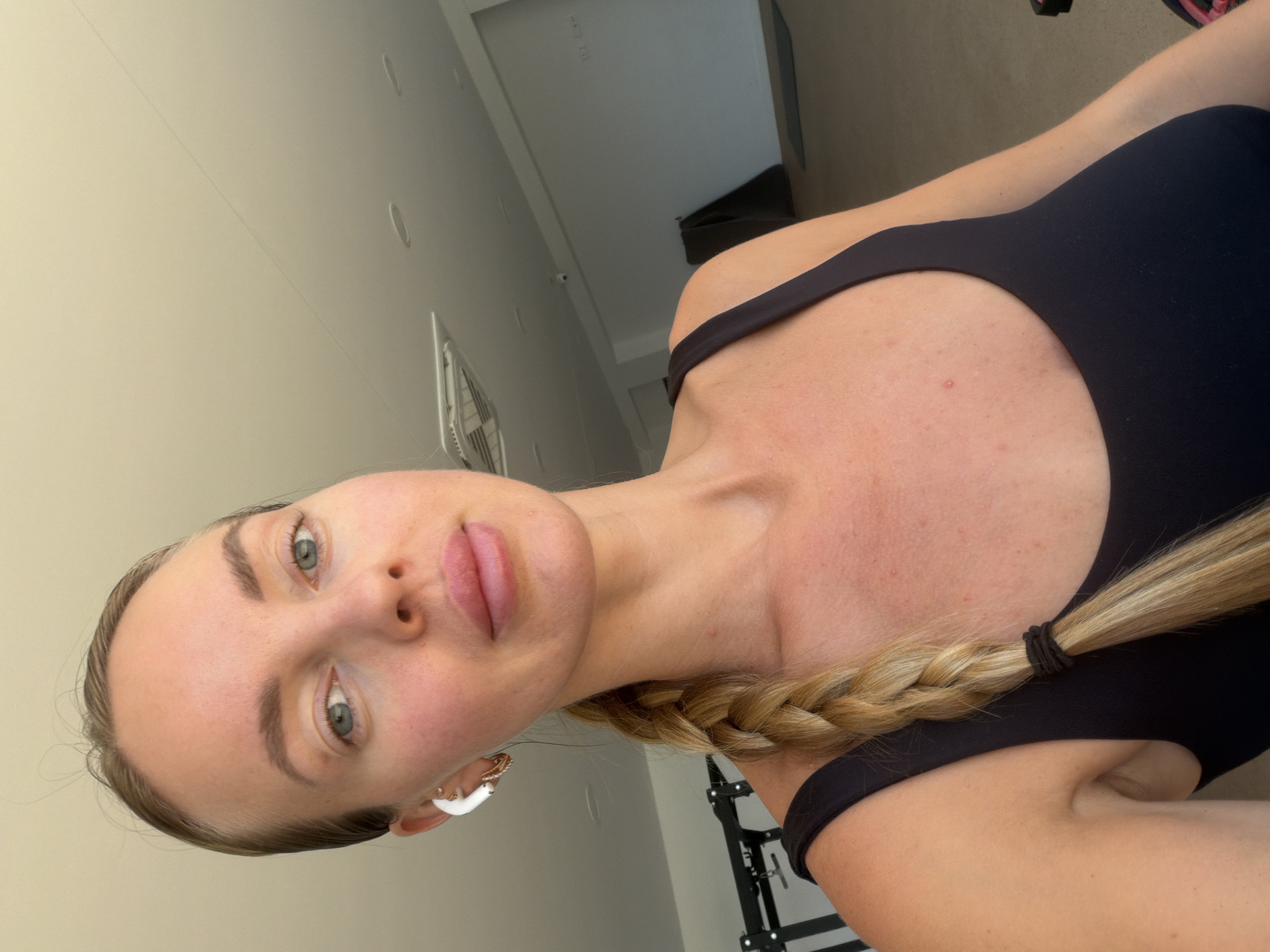 A close-up, over-the-shoulder, selfie-style photograph shows a fair-skinned woman with blonde hair in a braid. She is wearing a black athletic top and white earbuds. Her left ear is adorned with a white earbud and a hoop earring. The background is out of focus, but appears to be a gym with exercise equipment and a light-colored ceiling. The image was taken in Finestrat, Spain.