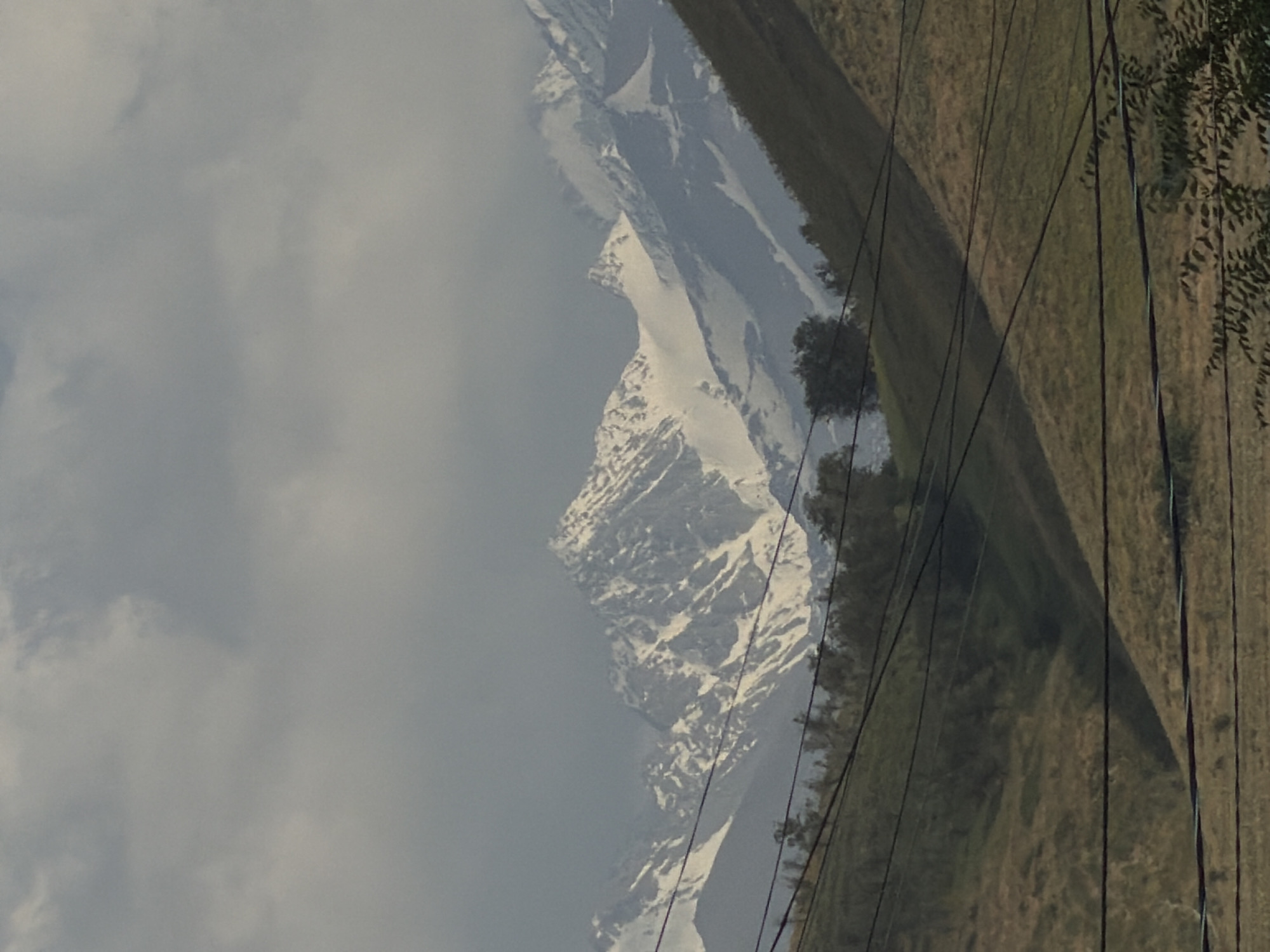A wide shot captures a mountainous landscape under a cloudy sky. The mountains in the background are snow-capped, with rugged peaks. In the foreground, rolling hills are covered in dry grass and sparse trees. Several power lines run horizontally across the frame, crisscrossing the view of the hills and mountains. The scene appears to be in a rural or undeveloped area.