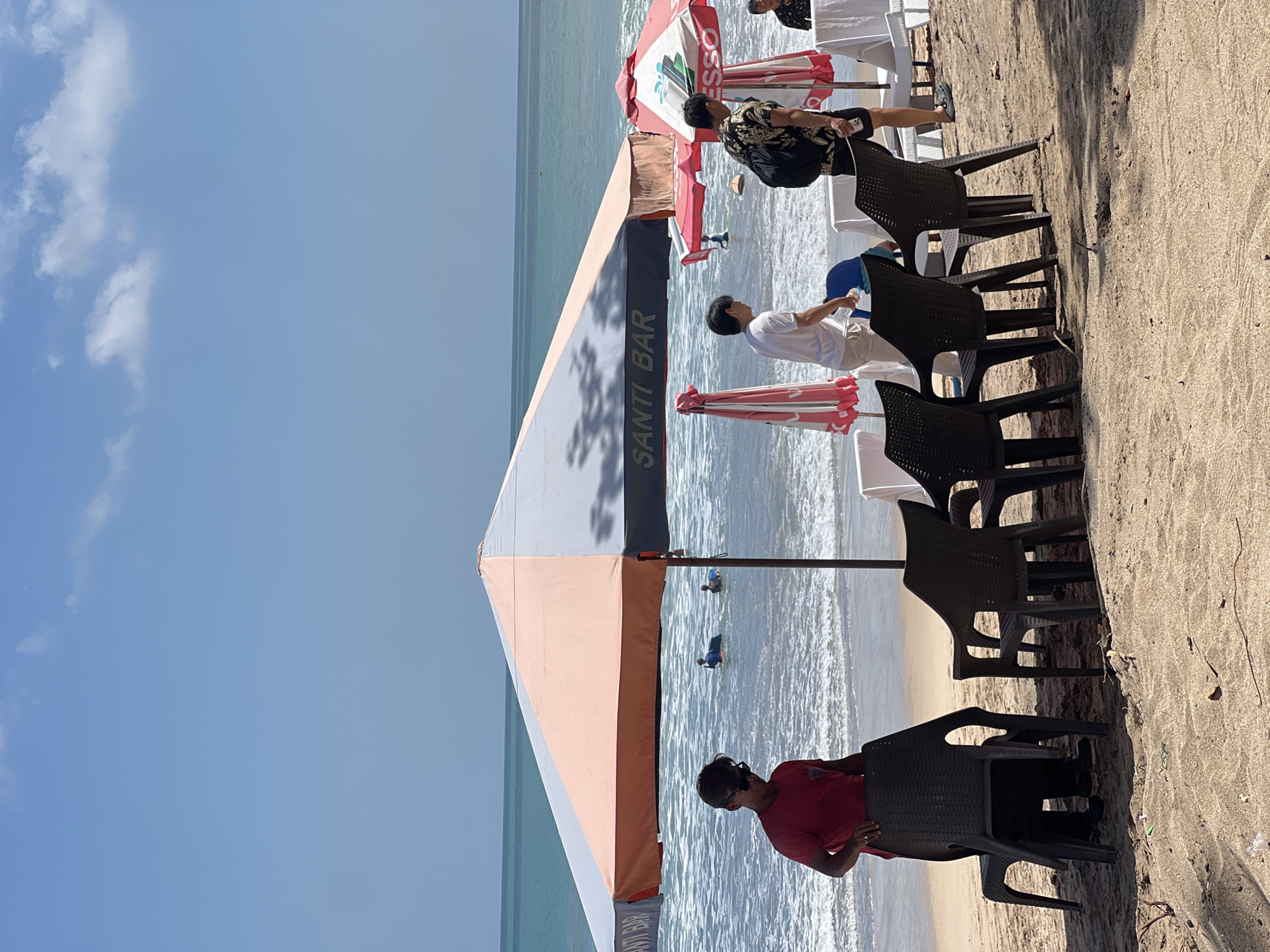 This is a photograph of Kuta Beach in Kuta, Indonesia, on a bright, sunny day. The foreground features a sandy beach with several dark plastic chairs and a large, multi-colored umbrella providing shade. In the background, the turquoise ocean is visible with gentle waves. Several people are in the water, some appearing to be on bodyboards. Two men are standing on the beach near the umbrella, one in a red shirt and sunglasses, and the other in a patterned shirt and shorts carrying a backpack. Two other men are seen walking towards the water, one in a white shirt and the other near a red umbrella. The overall atmosphere is that of a typical beach scene with people enjoying the sun and surf.