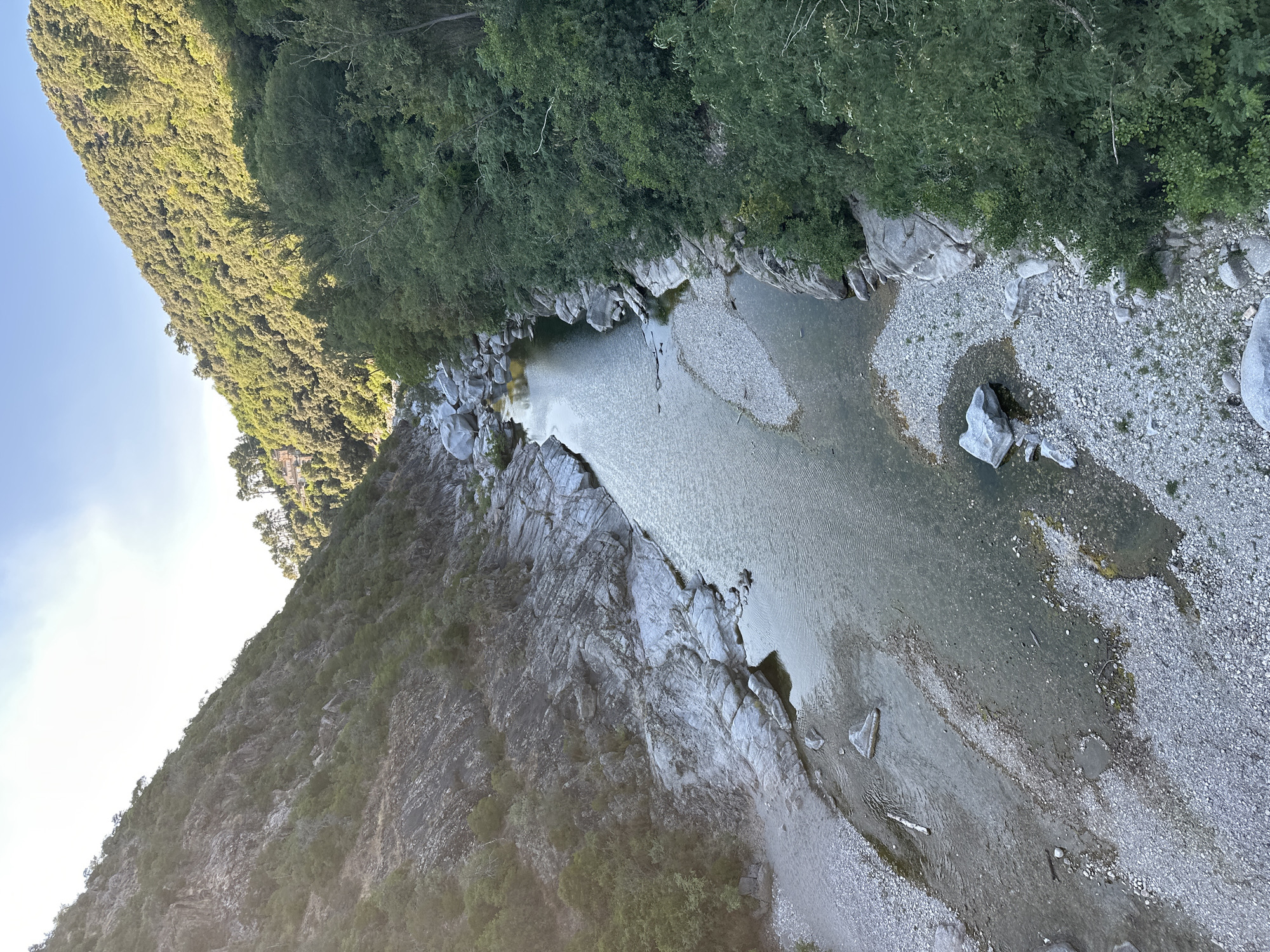 The image depicts a scenic view of a river flowing through a valley. The riverbanks are lined with rocks and trees, with the terrain rising into tree-covered hills on either side. The water is clear and reflects the light, with some areas of shallow riverbed visible. The scene appears tranquil and natural, with no human presence or activity evident. The lighting suggests it is either early morning or late afternoon, with the sun casting shadows and illuminating parts of the landscape. Based on the provided context, this image is likely of a location in Générargues, France.