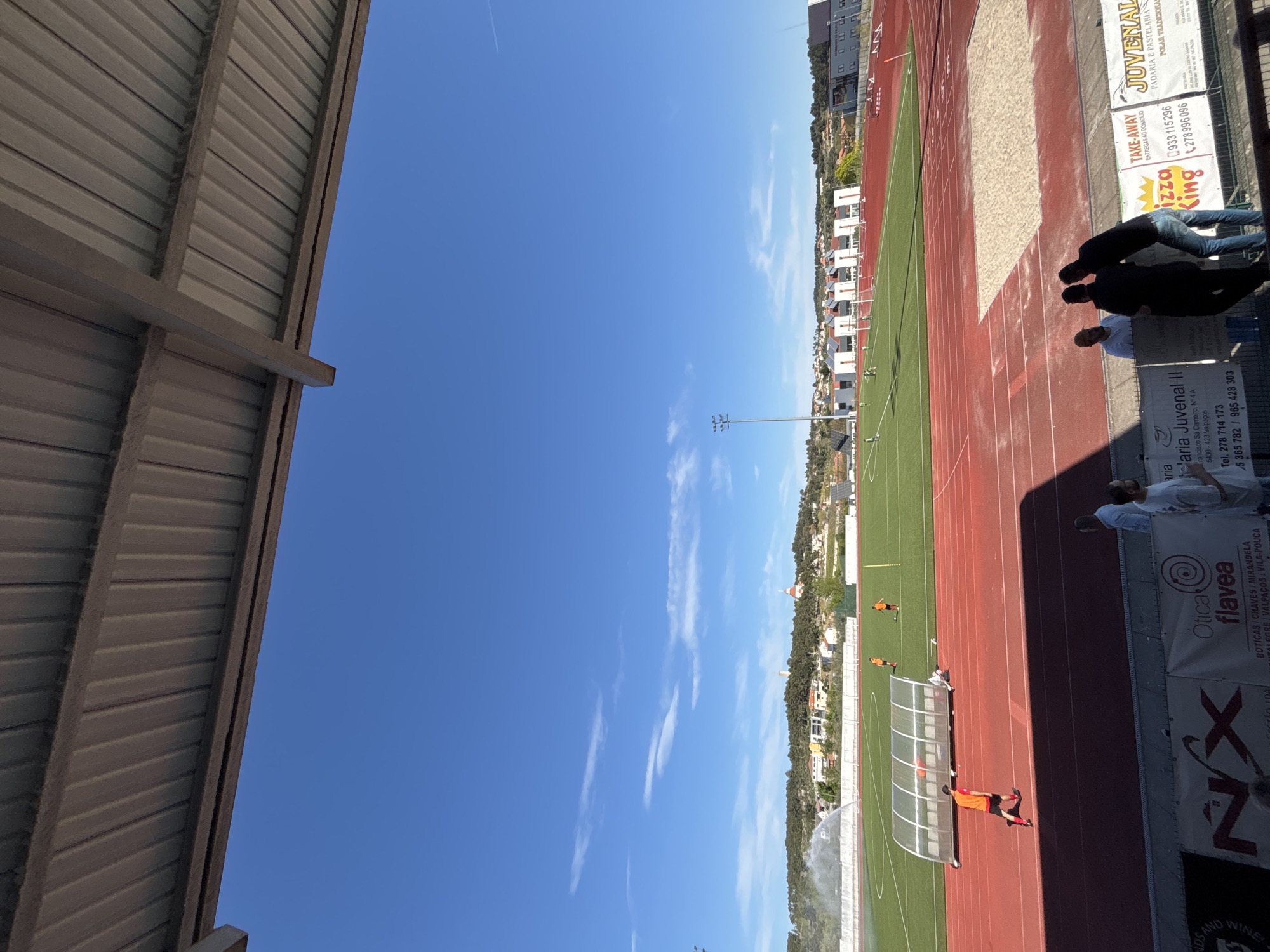 The image depicts a soccer field and running track in Valpaços, Portugal, adjacent to commercial banners and buildings. The sky is blue with wispy clouds. People are present near the banners, while soccer players are on the field, one near a sheltered area and another running. Sprinklers are watering the field. Buildings and trees dot the background landscape.