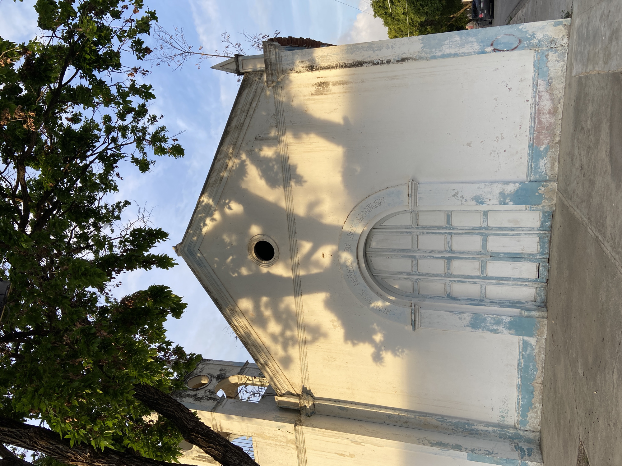 A medium-shot, eye-level, outdoor, vertical photograph captures a weathered, white-painted church facade with light blue accents at the base and around the entrance. The church, located in Juan Griego, Venezuela, features a prominent arched doorway with light blue, paneled double doors, set within a decorative white frame. Above the doorway, an inscription is visible in an arch. A circular window is positioned high on the gable. The building's facade shows signs of age, with peeling paint and stains. A bell tower, partially obscured by a tree on the left, extends upward with arched openings. The church is fronted by a concrete sidewalk. Lush green tree branches with leaves cast dappled shadows across the front of the church, particularly on the upper portion of the facade and the gable. The background reveals a clear blue sky with scattered clouds and the tops of trees. To the right of the church, a street with parked vehicles is visible, suggesting an urban or semi-urban setting. The lighting indicates it is likely late afternoon or early morning, with the sun casting warm light and long shadows.