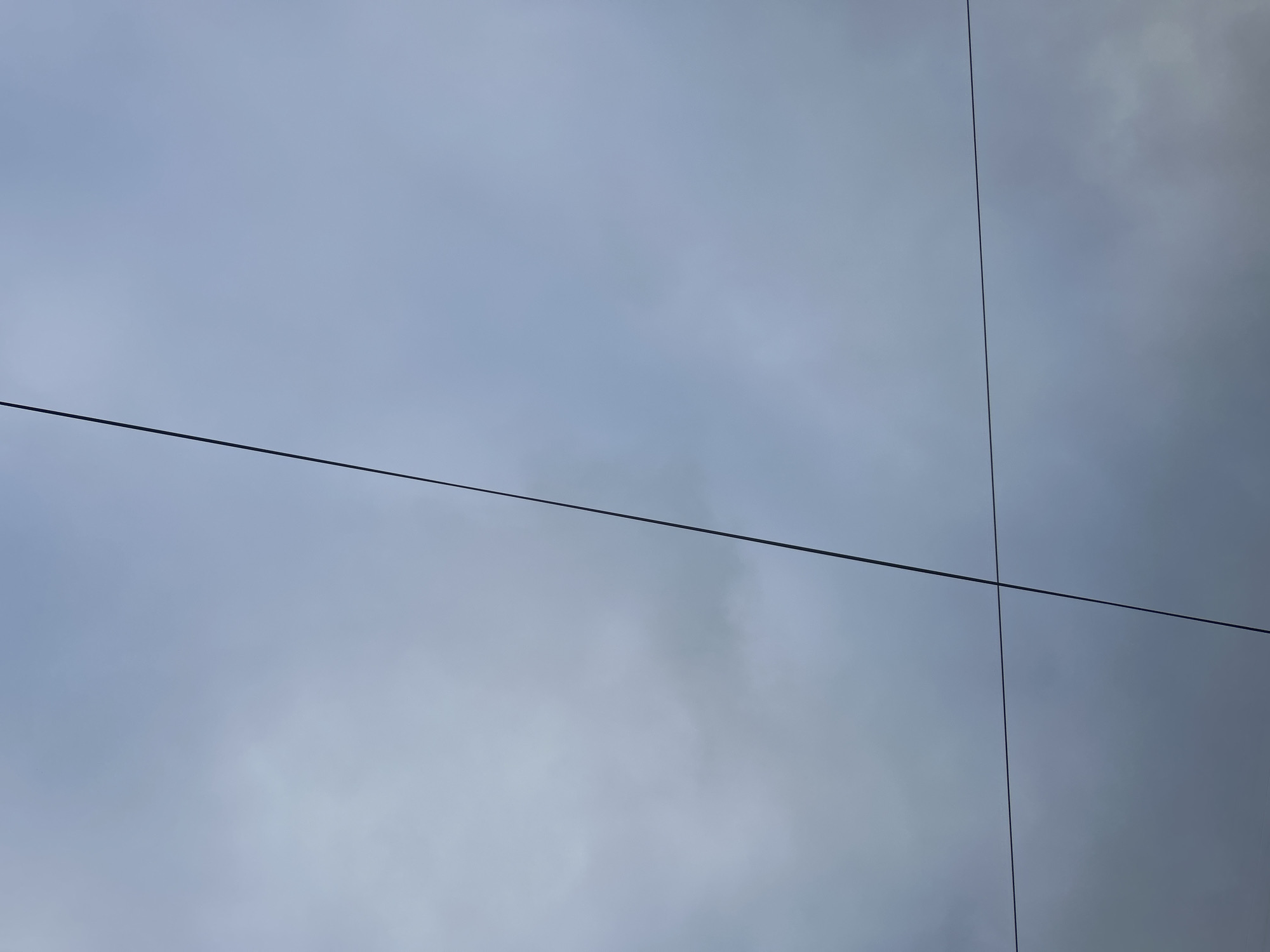 The image shows a cloudy sky reflected on a modern building facade in Câmara de Lobos, Portugal. Two thin black lines intersect, likely demarcating the edges of glass panels in the building's exterior. The sky is uniformly gray, with subtle variations in tone suggesting the presence of clouds.