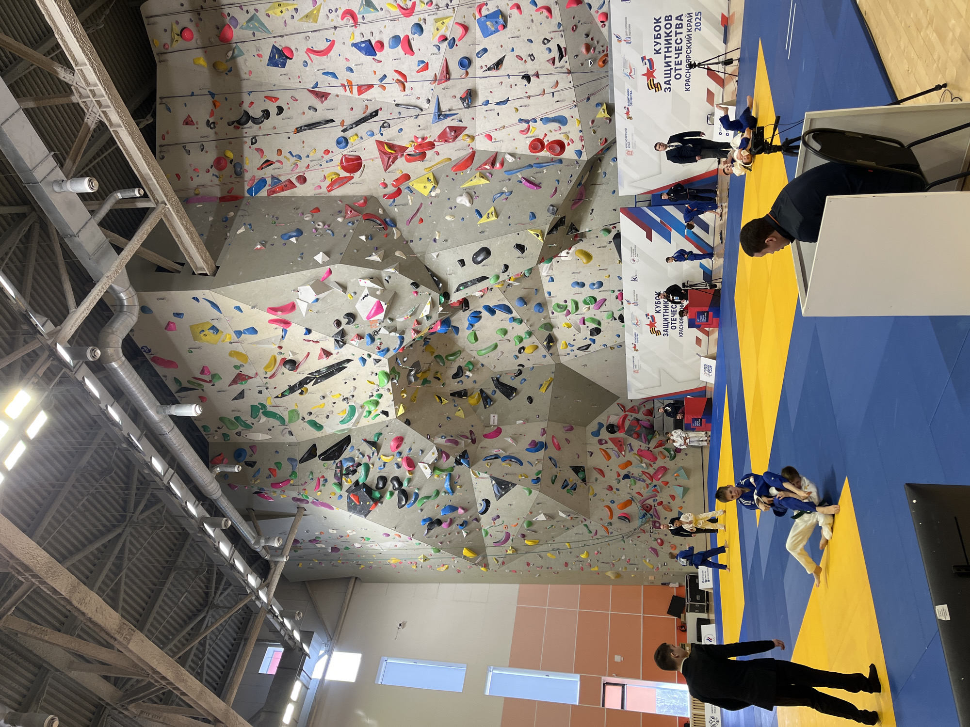 The image depicts an indoor sports facility, possibly a gymnasium or arena, in Krasnoyarsk, Russia. In the background is a climbing wall with a variety of colorful climbing holds affixed to a multi-faceted surface. In the foreground, a judo competition is in progress on a blue and yellow mat. Several judo participants wearing blue and white uniforms are engaged in grappling, with referees present. A few spectators are visible in the background behind the judo mat. There are banners that include Russian text and logos. The building has a high ceiling with visible support beams and lighting fixtures. Daylight enters through rectangular windows along one wall. A man in a suit and tie walks along the judo mat. A broadcast table is set up on the right. The image is slightly rotated counter-clockwise.