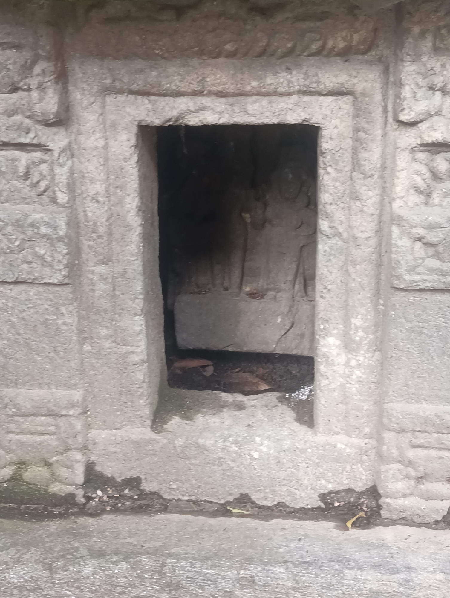 This is a close-up, eye-level, full outdoor shot of a recessed stone niche carved into a stone wall. The niche itself is dark, with some lighter-colored debris and a few dry leaves visible at the bottom. Within the niche, a carved relief is dimly visible, appearing to depict two standing figures. The stone surrounding the niche is rough-hewn and appears to be weathered. The wall has decorative elements on either side of the niche, with horizontal lines and some textured patterns. The ground in front of the wall is also stony and appears to have some damp patches and debris. The image is not in focus. There is no indication that this location is in Gojra, India.