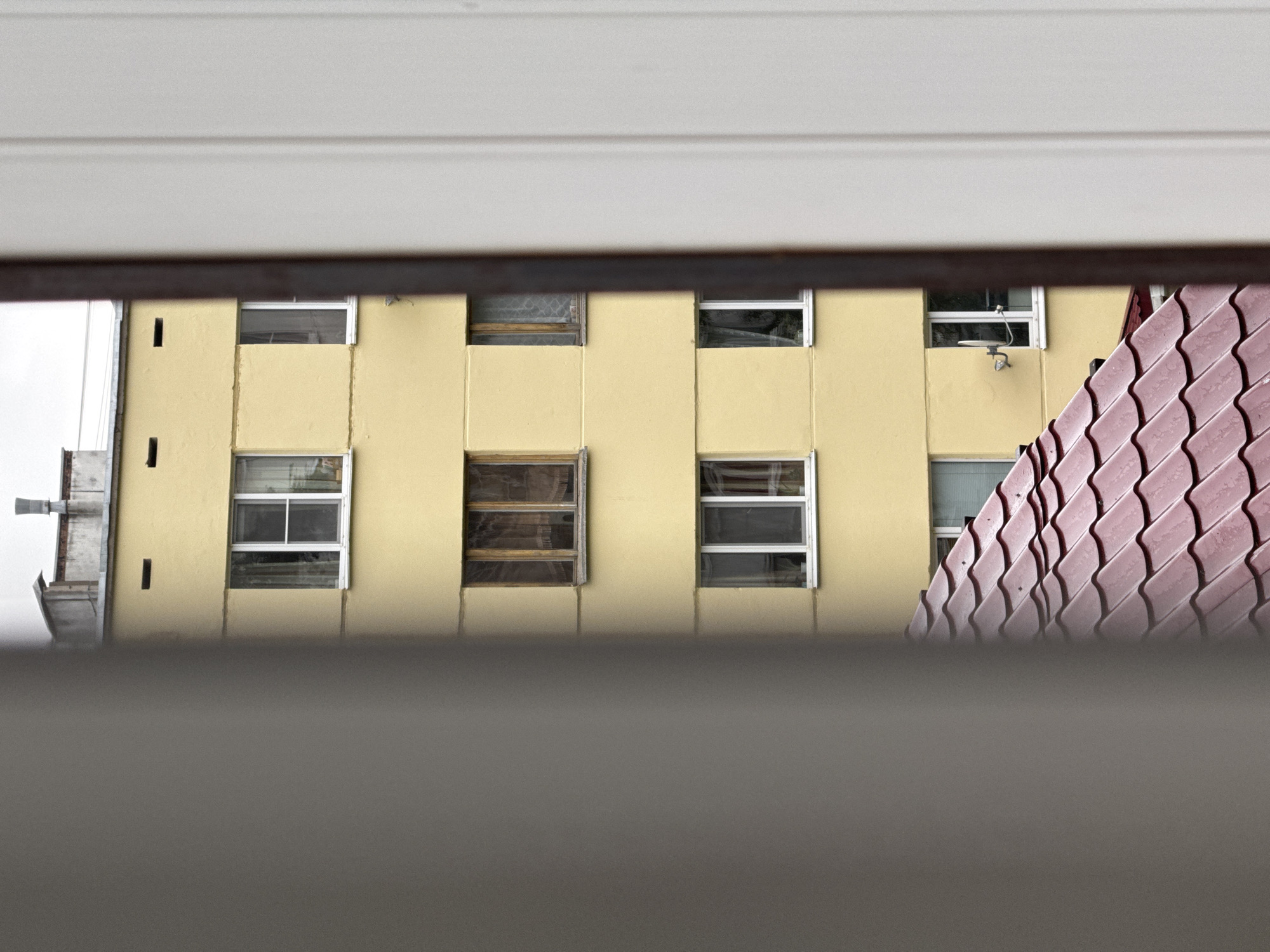 The image shows a view through a window of a pale yellow building. The building has several windows, some of which are visible. There is also a portion of a reddish, wavy tiled roof on the right side of the image. On the left side, there is a metal structure that appears to be part of the building's exterior or some sort of equipment. The overall scene suggests an urban or suburban environment. The lighting appears to be overcast, possibly indicating a cloudy day or early morning/late afternoon. There are no people or animals visible in the image. The focus is on the architectural details of the building and the roof. No text is visible in the image.