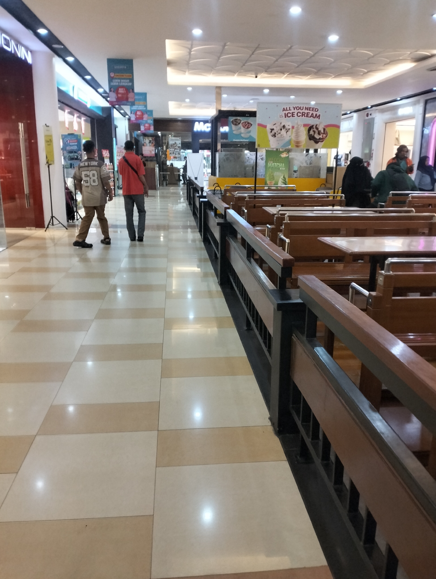 The image depicts a corridor within a shopping mall in Semarang, Indonesia. The floor is tiled with a pattern of beige and cream squares. To the left, a shopfront with red and white signage is visible. Further down the corridor, two men are walking away from the camera. The man on the left wears a light brown t-shirt with the number "58" and "NEW YORK" printed on it, and brown pants. The man on the right wears a red t-shirt and grey pants. On the right side of the corridor, a seating area with wooden benches and tables is set up. A banner advertising "ALL YOU NEED is ICE CREAM" is displayed above the seating area. The ceiling is white and features recessed lighting. Several people are visible in the background, some seated in the dining area, and others walking further down the corridor. The overall atmosphere suggests a public indoor space within a commercial establishment.