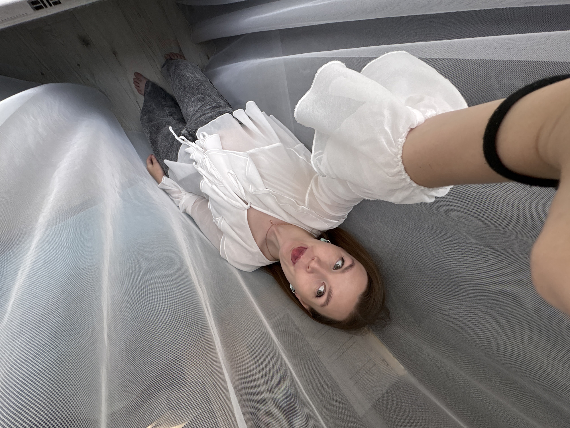 A young woman with brown hair is lying down, partially covered by sheer white fabric. She is wearing a white ruffled blouse and gray pants. Her right arm is extended, holding a phone to take a selfie. She is looking at the camera with an open mouth and her tongue sticking out slightly. The background appears to be a room with wooden floorboards and sheer curtains. The image does not contain information about the city of Izhevsk, Russia.