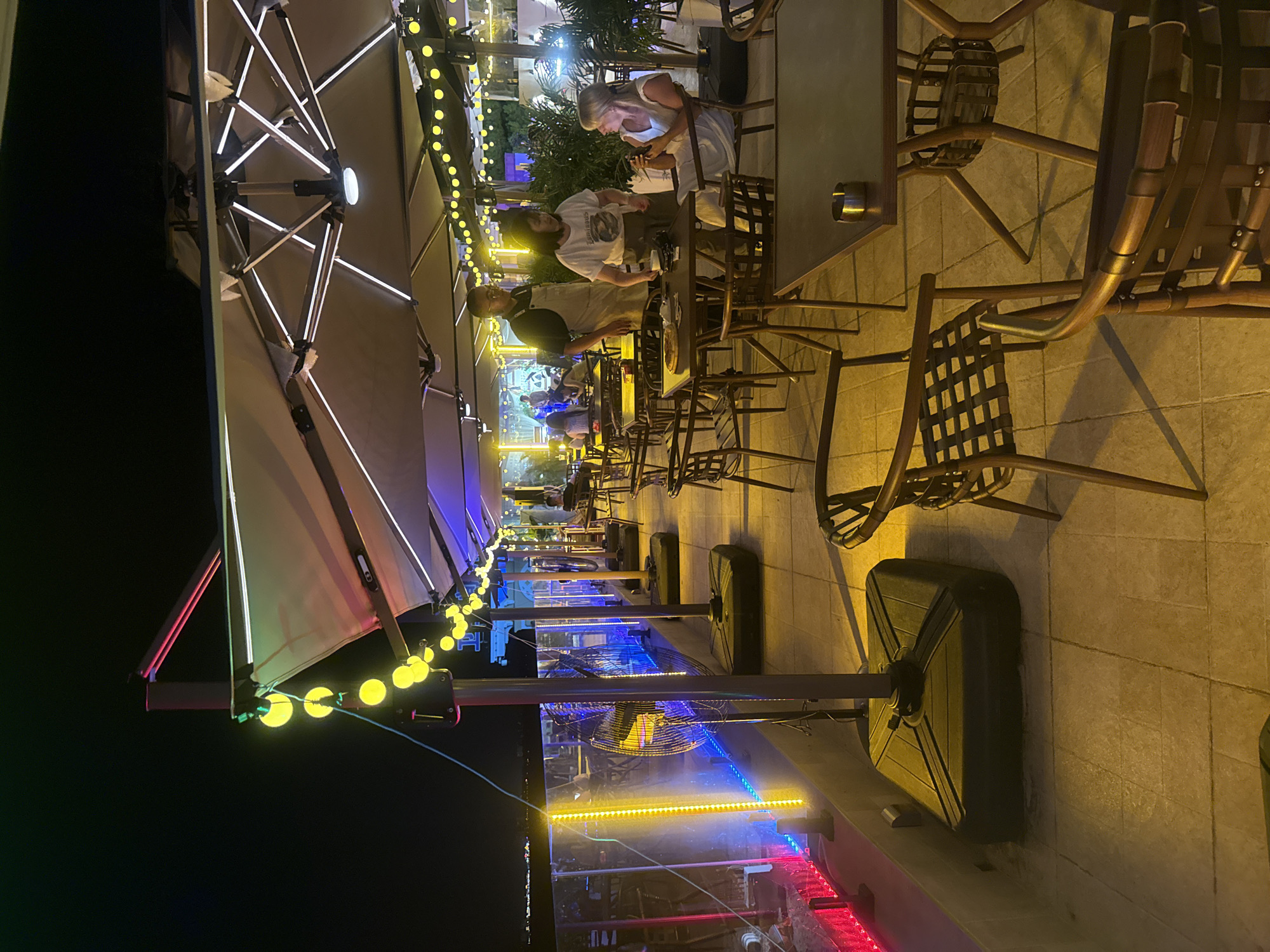 An outdoor dining area at night, likely a restaurant or cafe, is depicted in this image. The scene is illuminated by colorful string lights and under-canopy lighting, casting a warm glow on the tiled floor and outdoor furniture. Several large umbrellas provide shade. In the foreground, a woman in a white dress is seated at a table, holding a phone. To her left, a server in a light-colored apron and a woman in a t-shirt are engaged in conversation. Other tables and chairs are visible, some occupied by diners in the background. Large industrial fans stand near the edge of the patio, and decorative lighting accents the railing. The distant city lights suggest an urban setting, and the presence of palm trees hints at a warm climate. While the provided information indicates the location as Qinhuangdao, China, this is not visually ascertainable from the image itself.