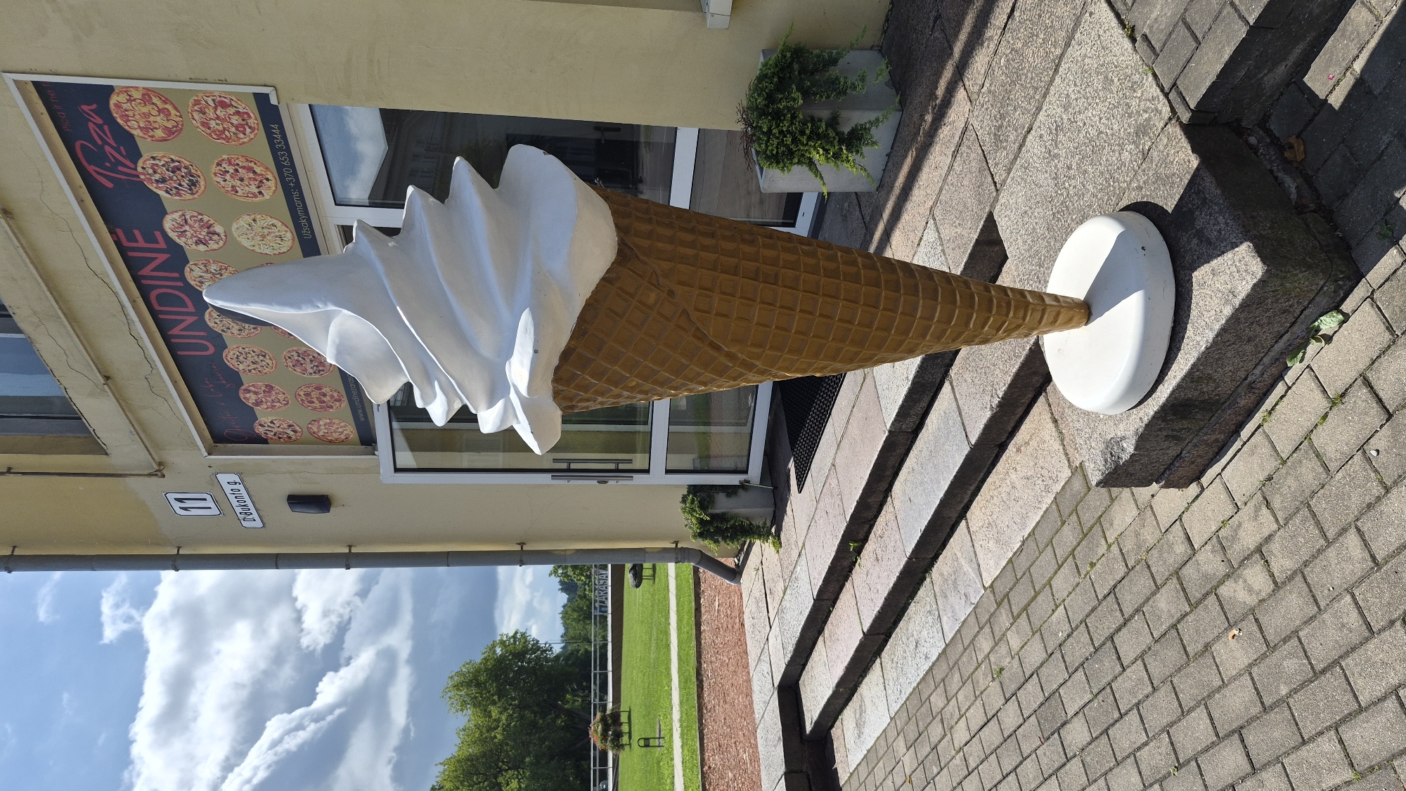The image captures a large, artificial ice cream cone sculpture standing on a granite base in front of a building. The building's facade is painted a light yellow, and above the entrance is a sign that reads "UNDINE Pizza" and displays multiple pizza images. A street sign on the building shows "11" and "D-Bukonto g.", indicating the location is Bukonto Street, number 11, in Zarasai, Lithuania. The sculpture is white and swirled at the top, resembling soft-serve ice cream, and is placed on a white circular stand. The cone itself is a golden-brown color with a waffle pattern. To the left of the entrance, a set of stone steps leads up to the building, with a paved walkway in the foreground. In the background, a green lawn with some trees and benches is visible under a partly cloudy sky. Two potted plants are situated near the entrance, one on each side. The scene appears to be an outdoor commercial area.
